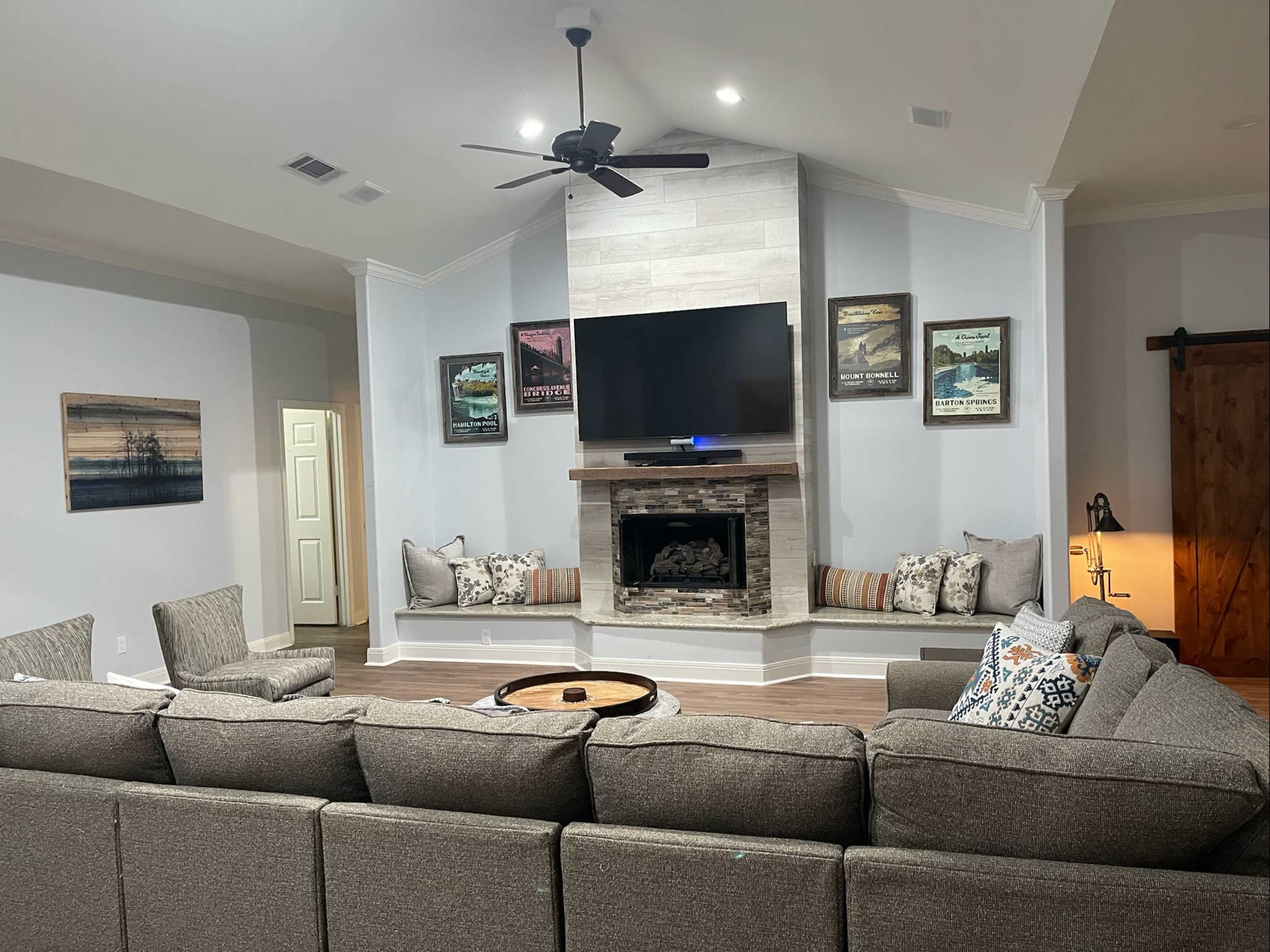 A spacious living room features a sectional sofa facing a stone fireplace with a mounted television above it, framed artwork on the walls, and cozy seating areas on either side of the fireplace.
