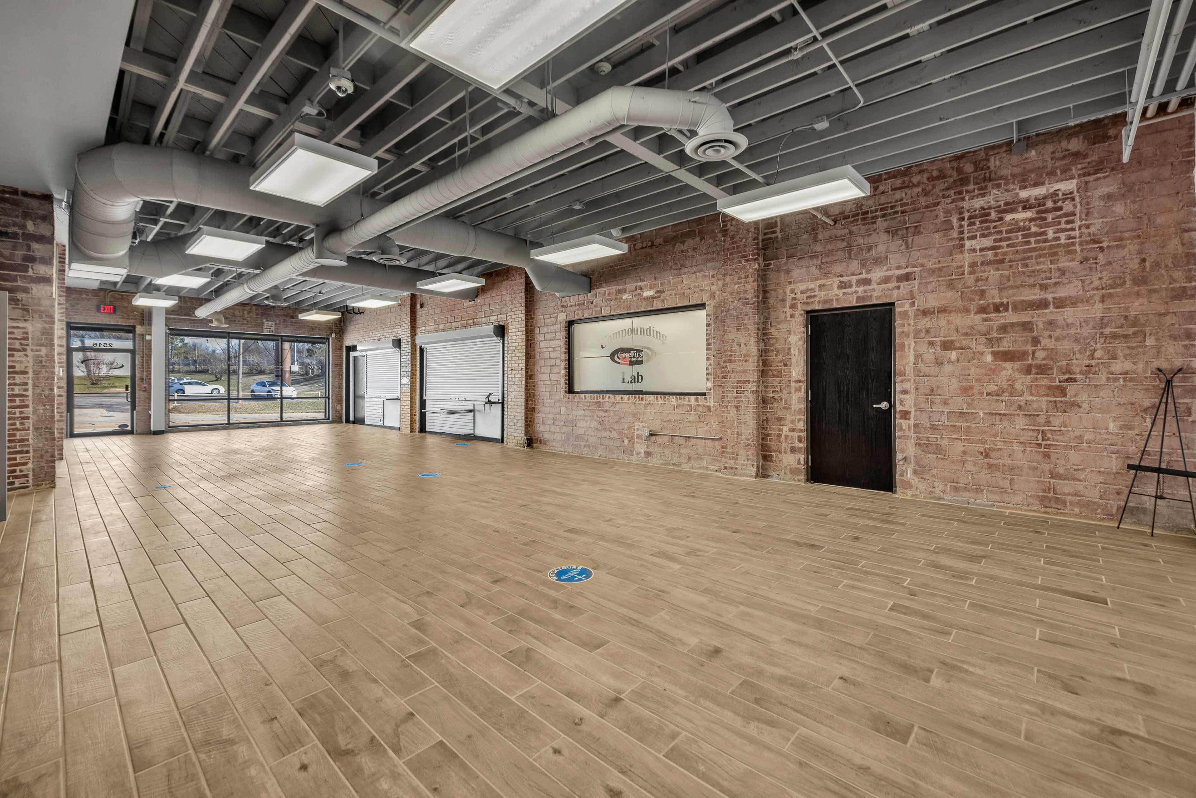 The image shows an empty interior space with wooden flooring, exposed brick walls, and large windows providing natural light.