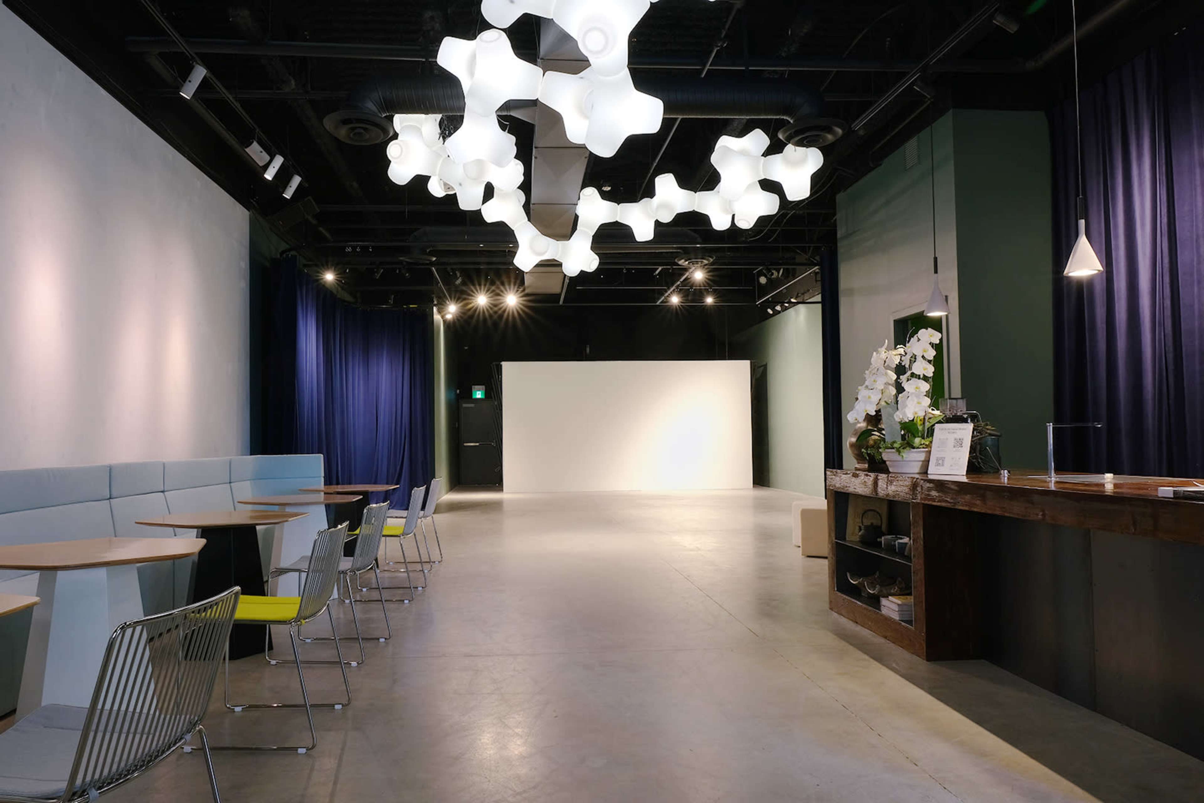 A modern, open space with tables and chairs on one side, a counter with a decorative plant, and a large blank wall in the background, illuminated by unique overhead lighting fixtures.