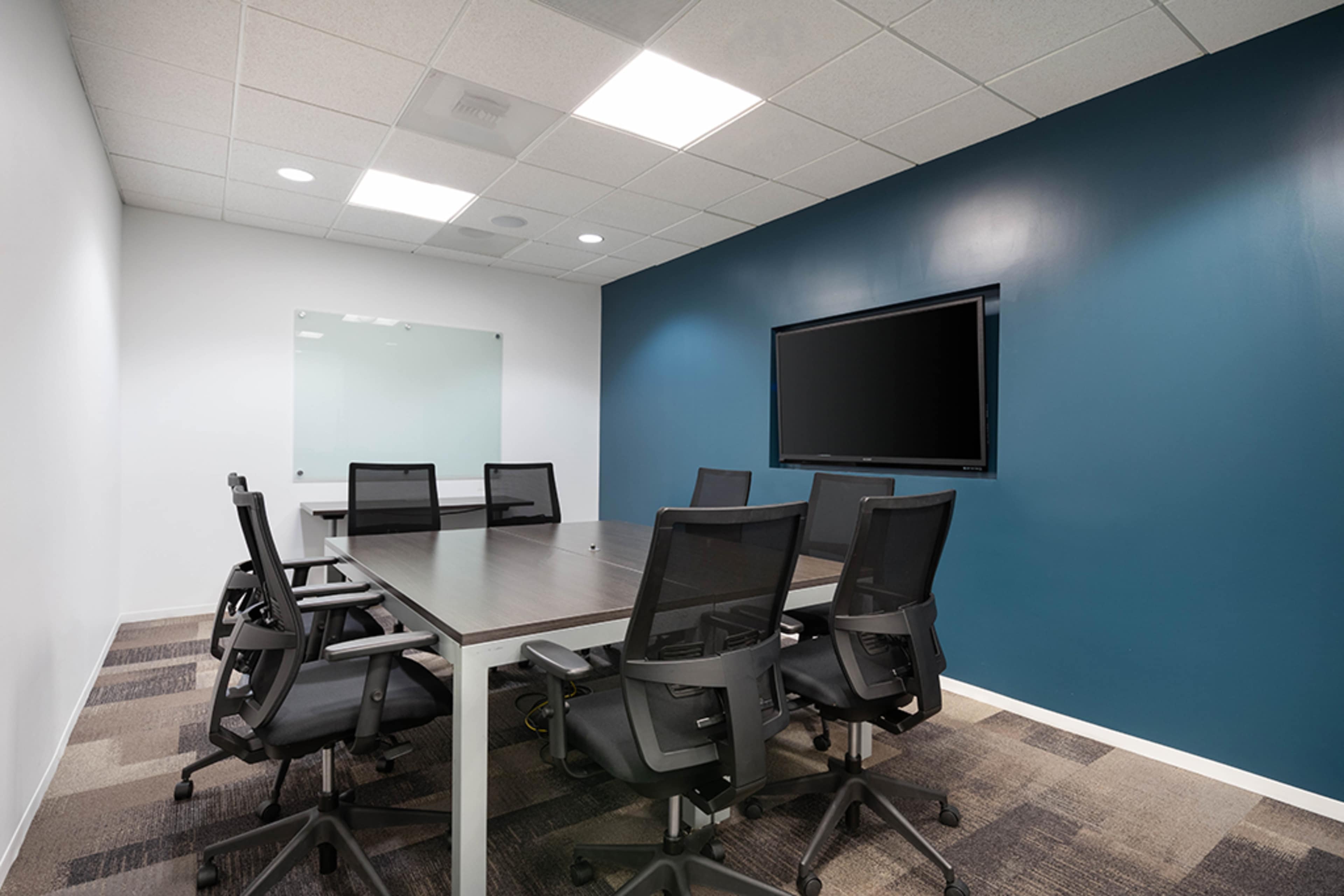 A modern conference room features a rectangular table surrounded by ergonomic chairs, a large screen on the wall, and a glass whiteboard.