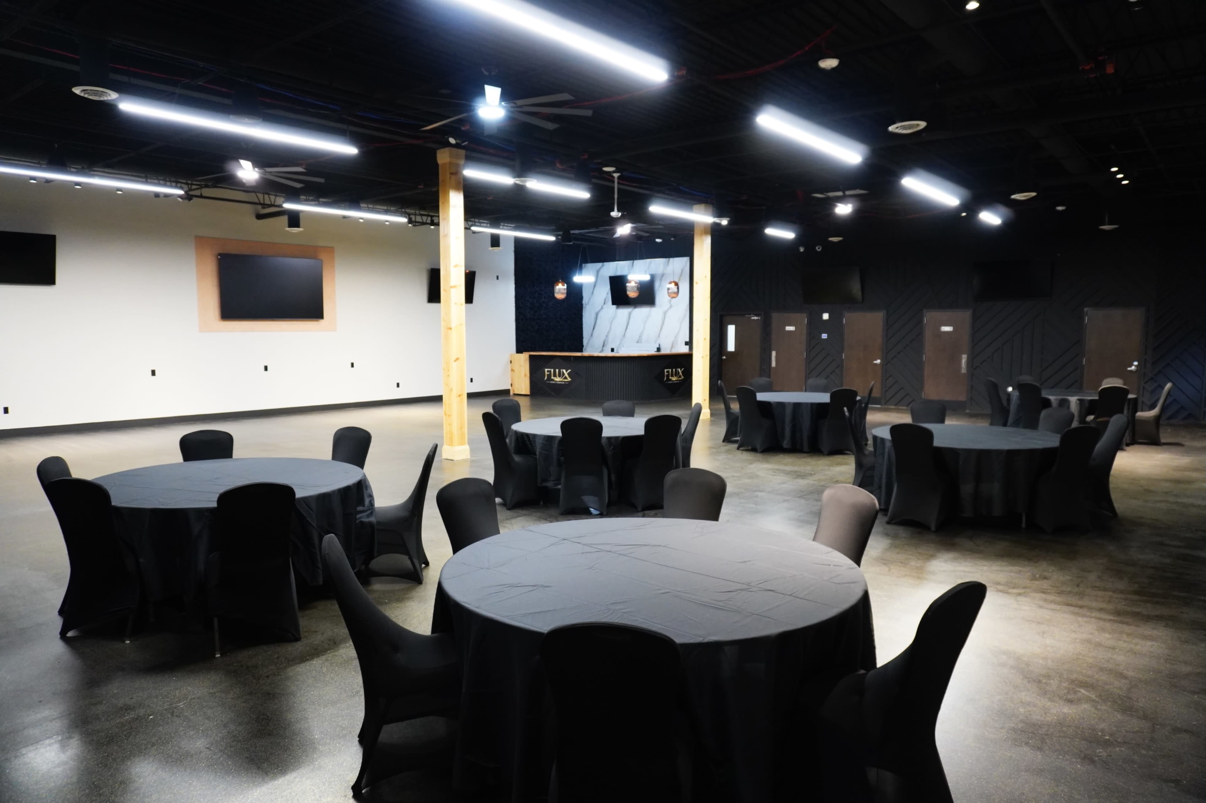 The image shows a large event space featuring several round tables with black tablecloths arranged in a well-lit area, with a bar and screen at one side.