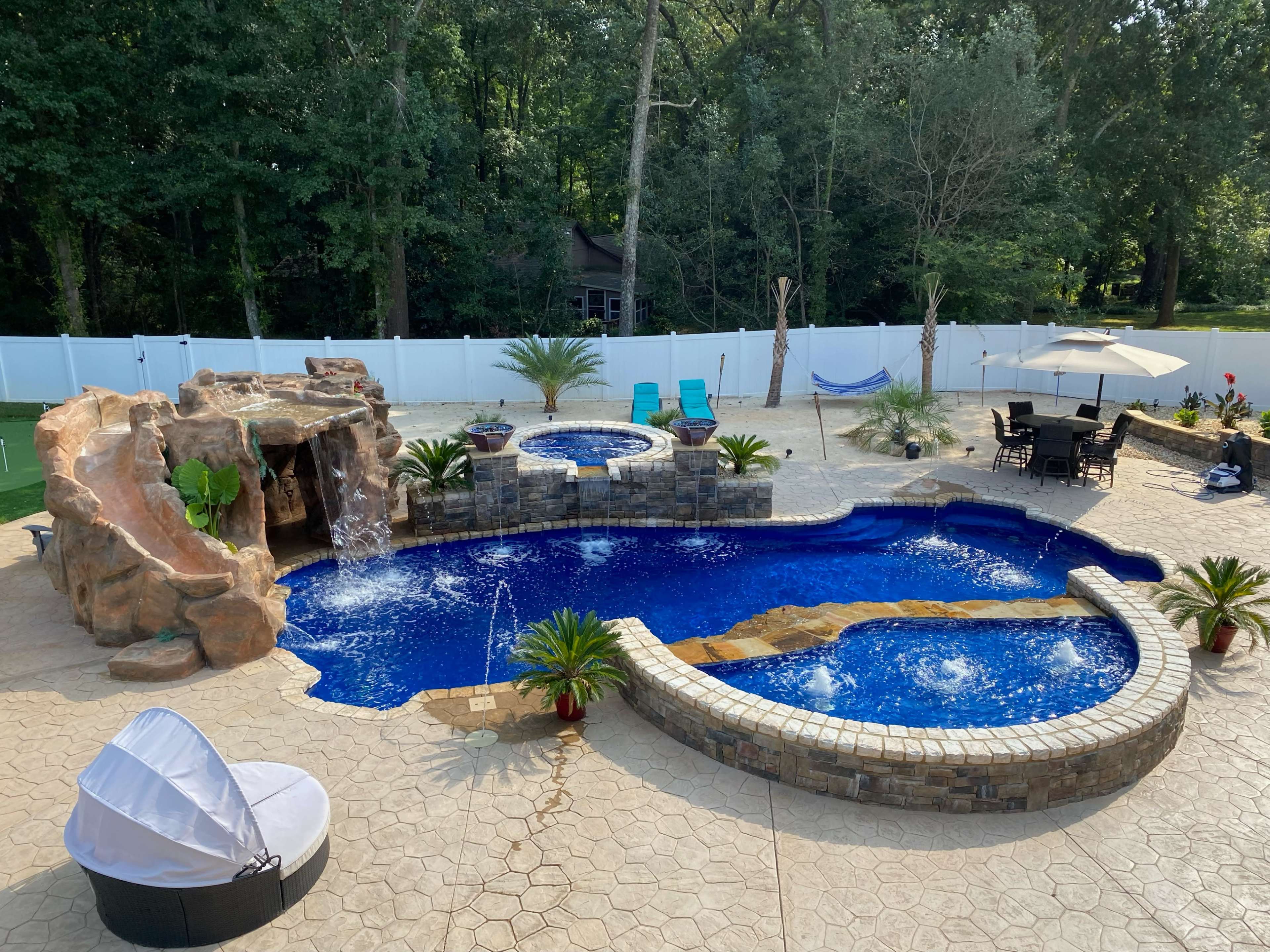 A backyard pool area featuring a large, curved swimming pool with a rock waterfall, a hot tub, lounge chairs, and landscaped greenery.