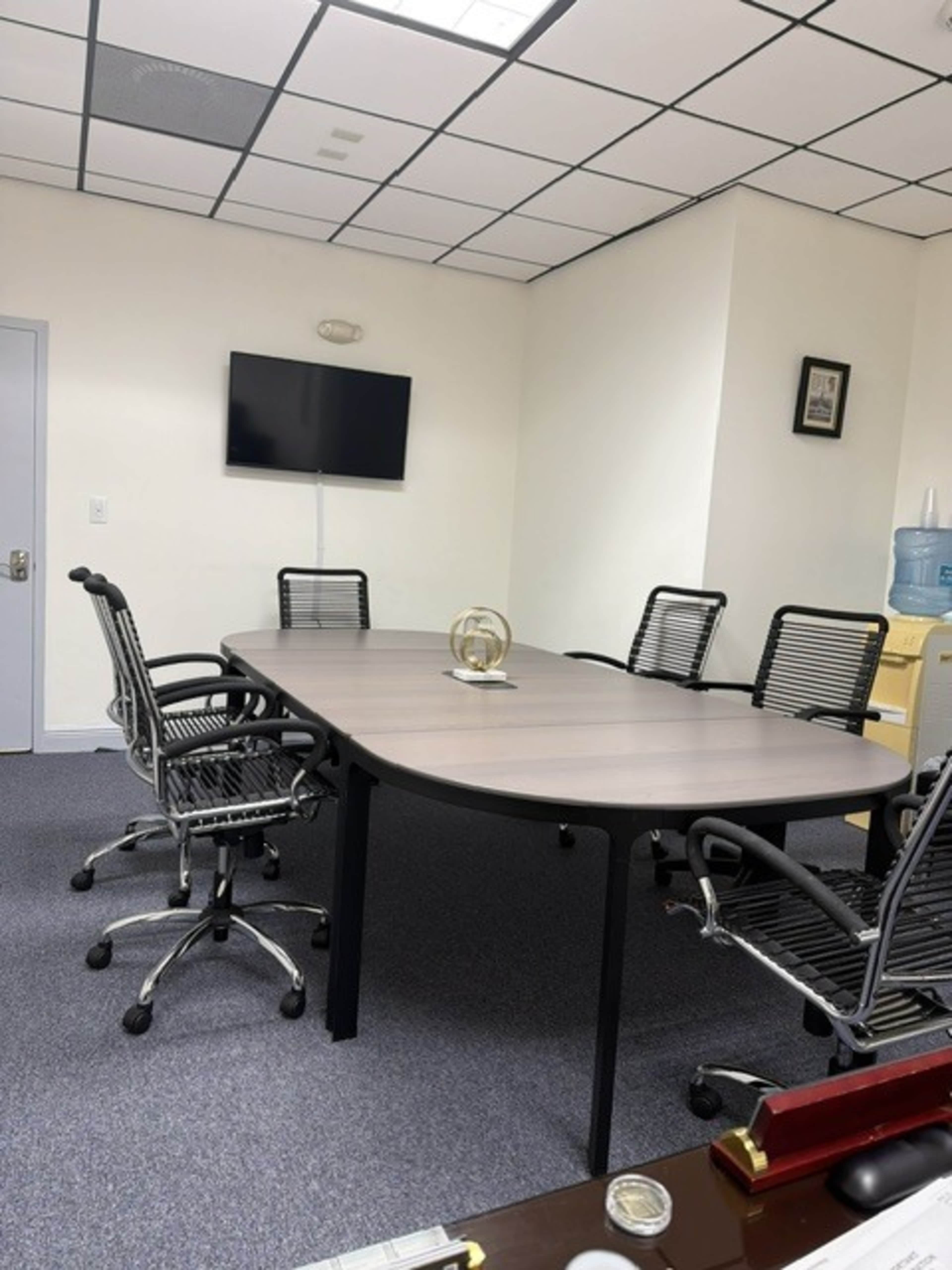 The image shows a conference room with a large table surrounded by several office chairs, a wall-mounted television, and a water dispenser in the background.