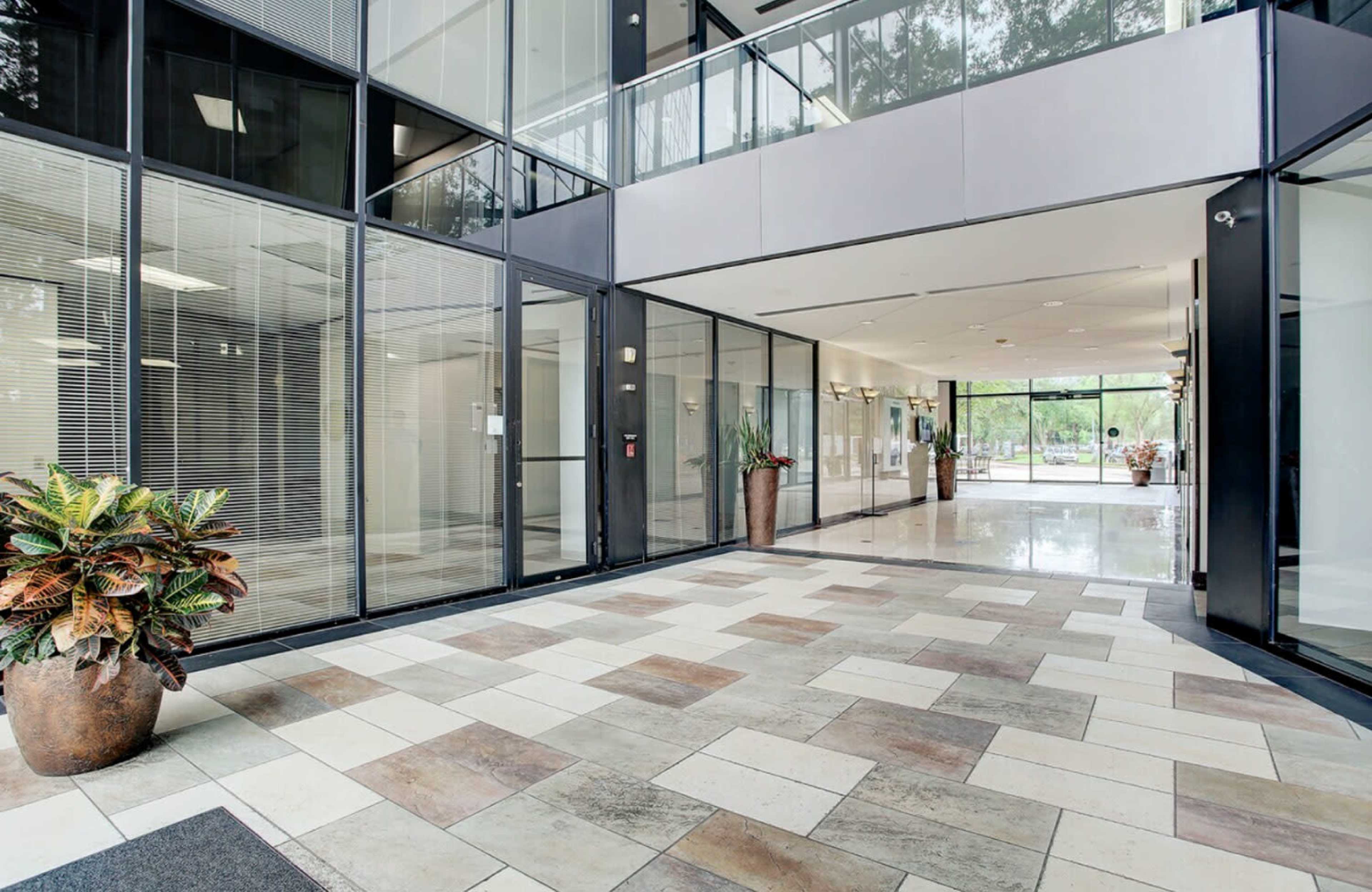 The image shows a modern lobby with large glass panels, polished tile flooring, and potted plants near the entrance.