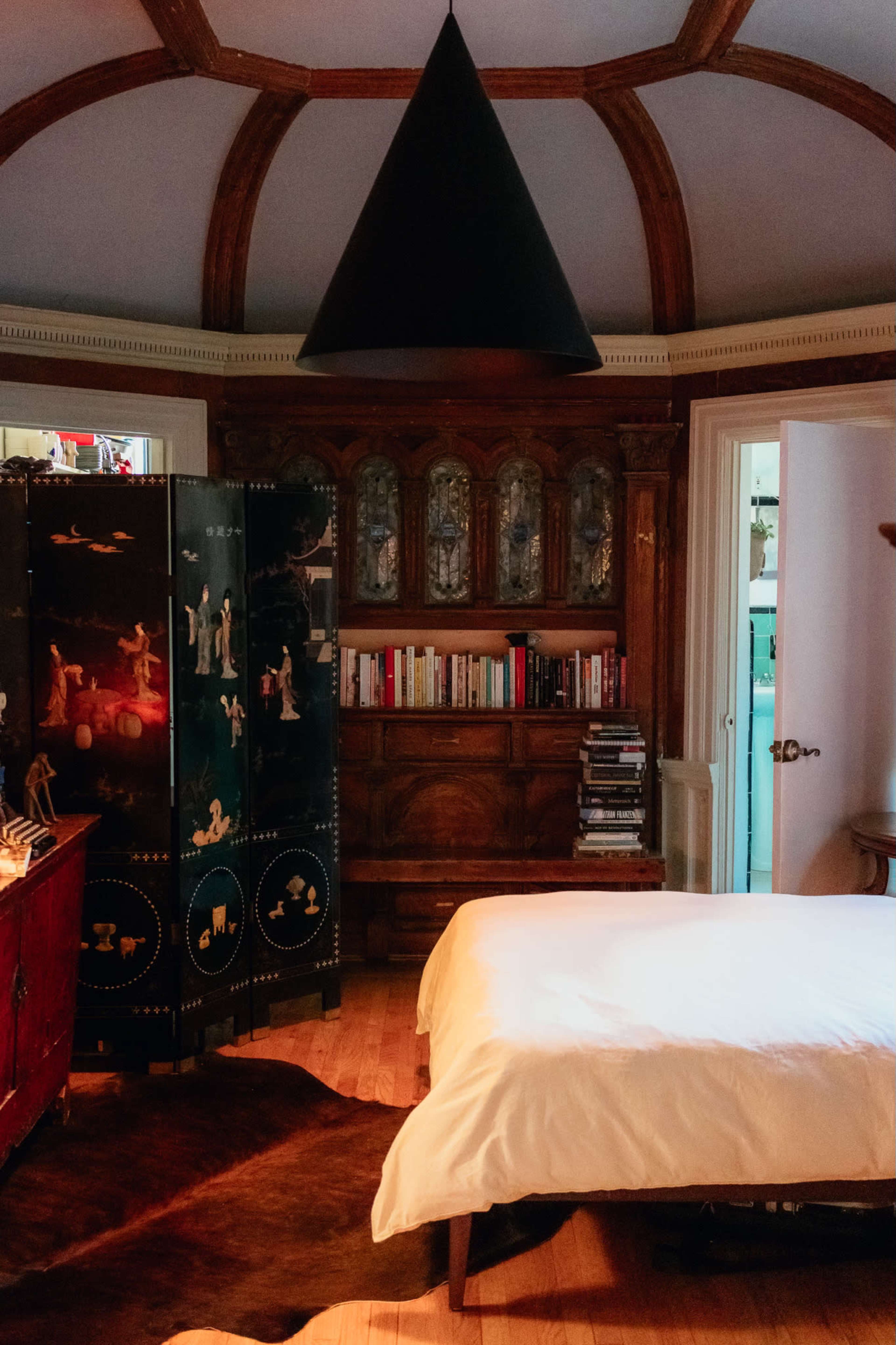 A cozy bedroom features a white bed, an ornate wooden bookshelf, and a distinctive black cone-shaped light fixture hanging from the ceiling.