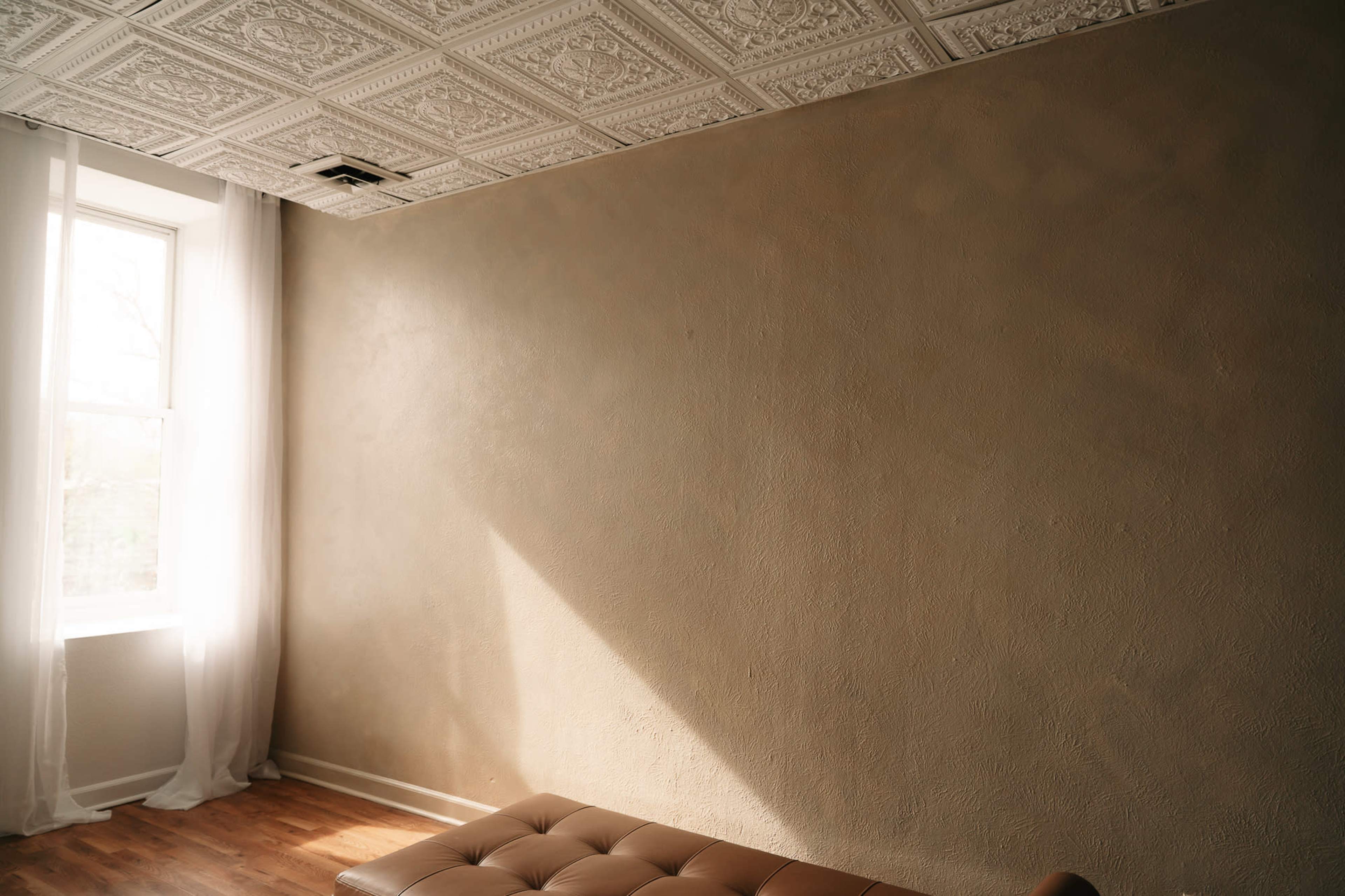 The image shows a room with a textured brown wall, a large window letting in natural light, and a brown tufted leather bench.