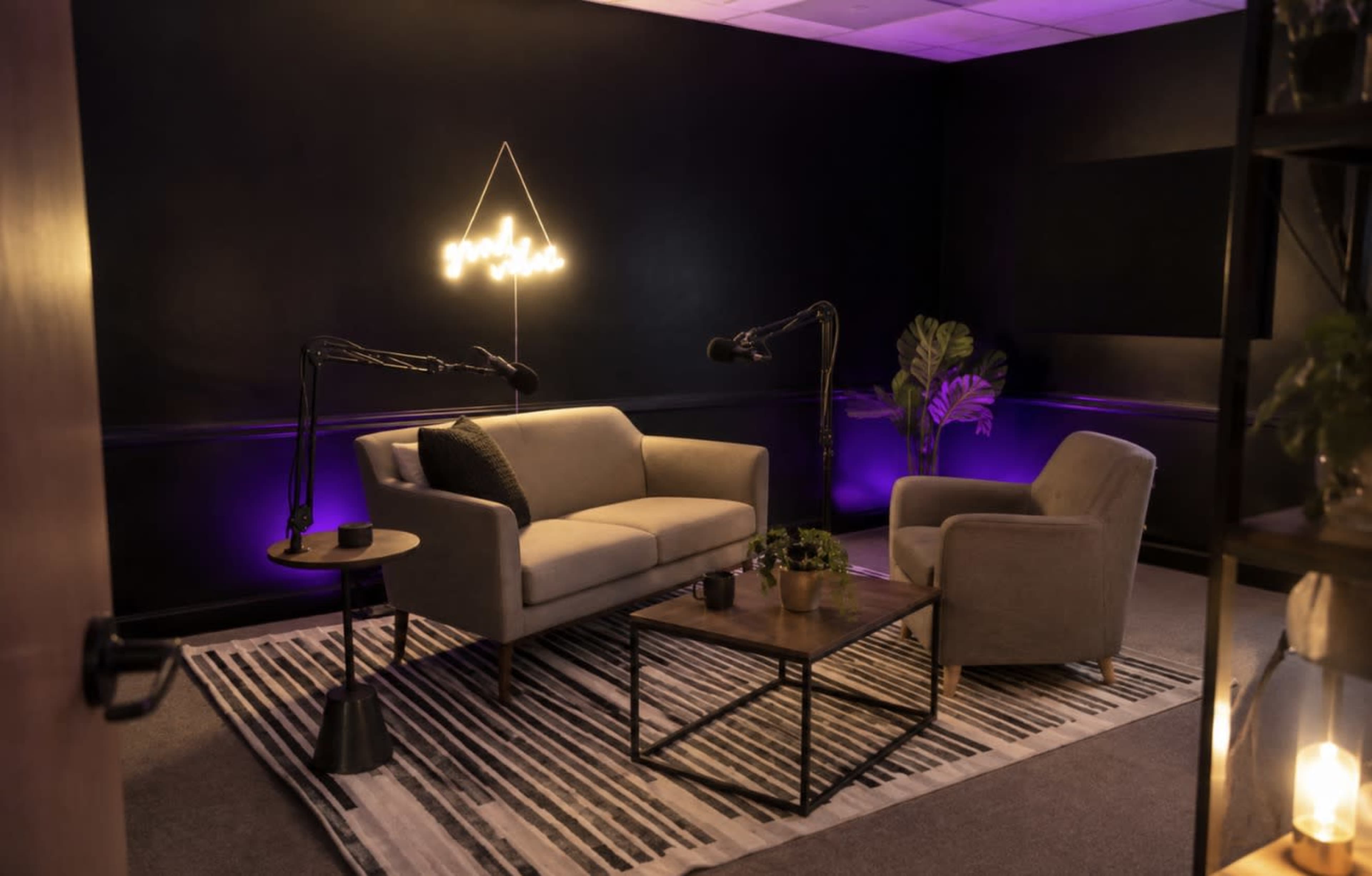 The image shows a modern lounge space with a gray sofa, an armchair, a coffee table, and neon lighting on a dark wall, creating a cozy atmosphere ideal for conversation or recording.