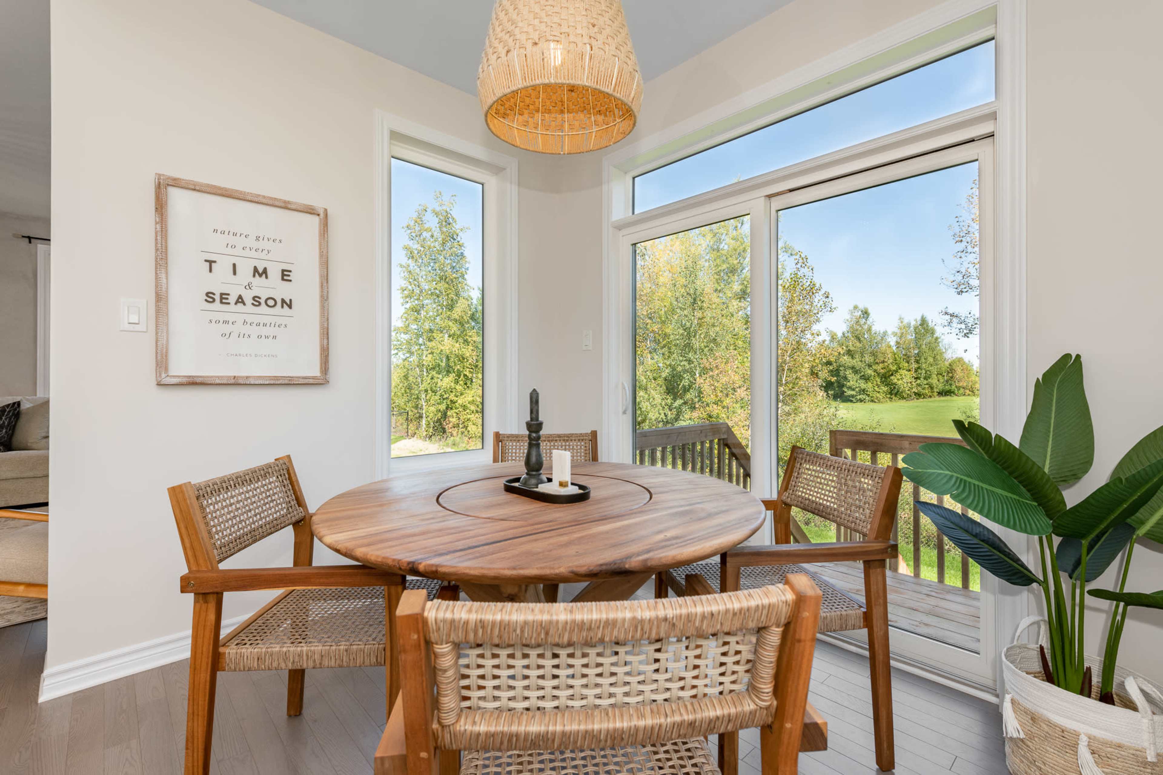A round wooden dining table with four chairs is set near large windows overlooking a green landscape.