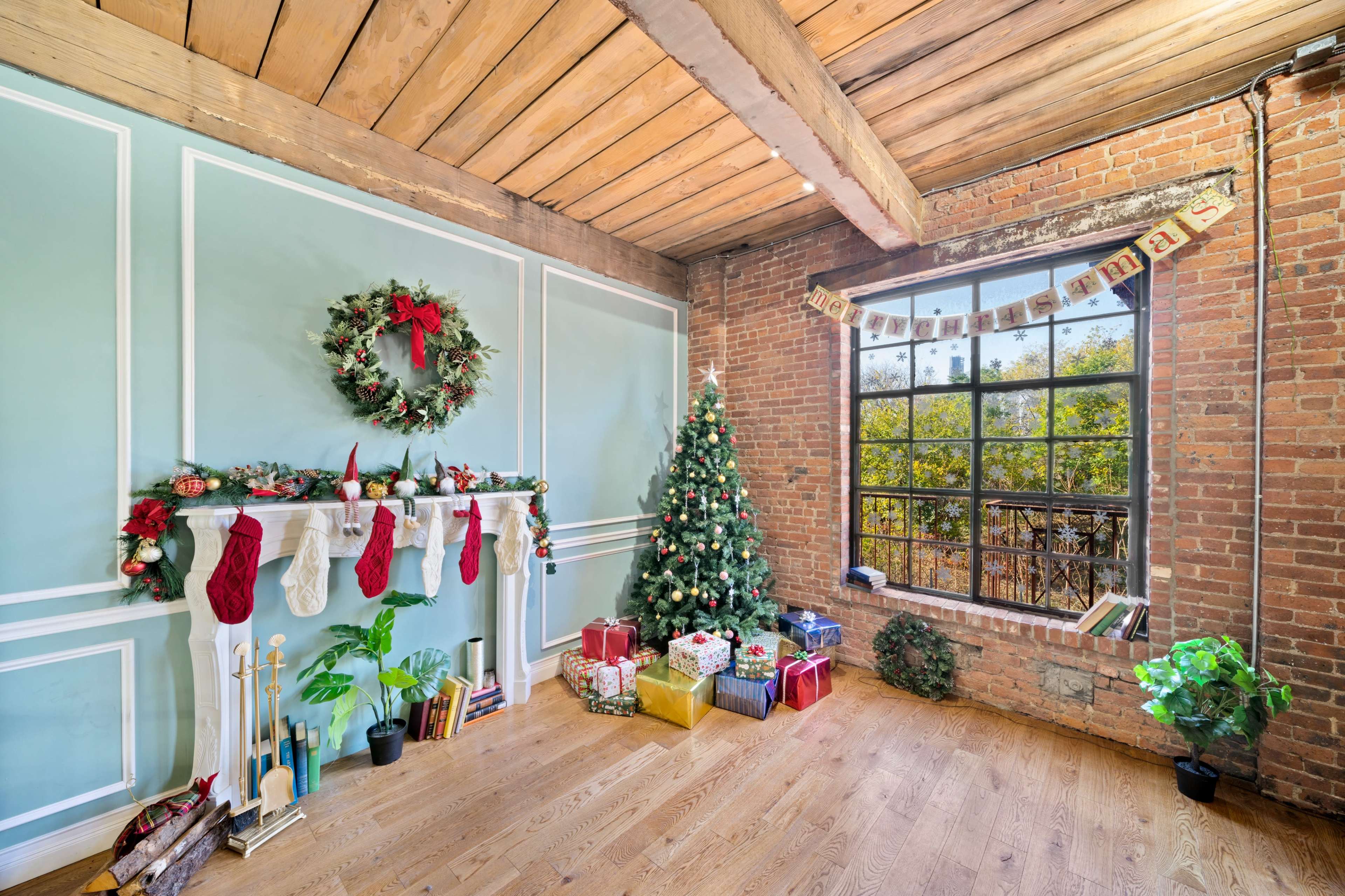 A decorated living room features a Christmas tree, stockings hung on a white mantel, and wrapped gifts on the wooden floor beside a large window.