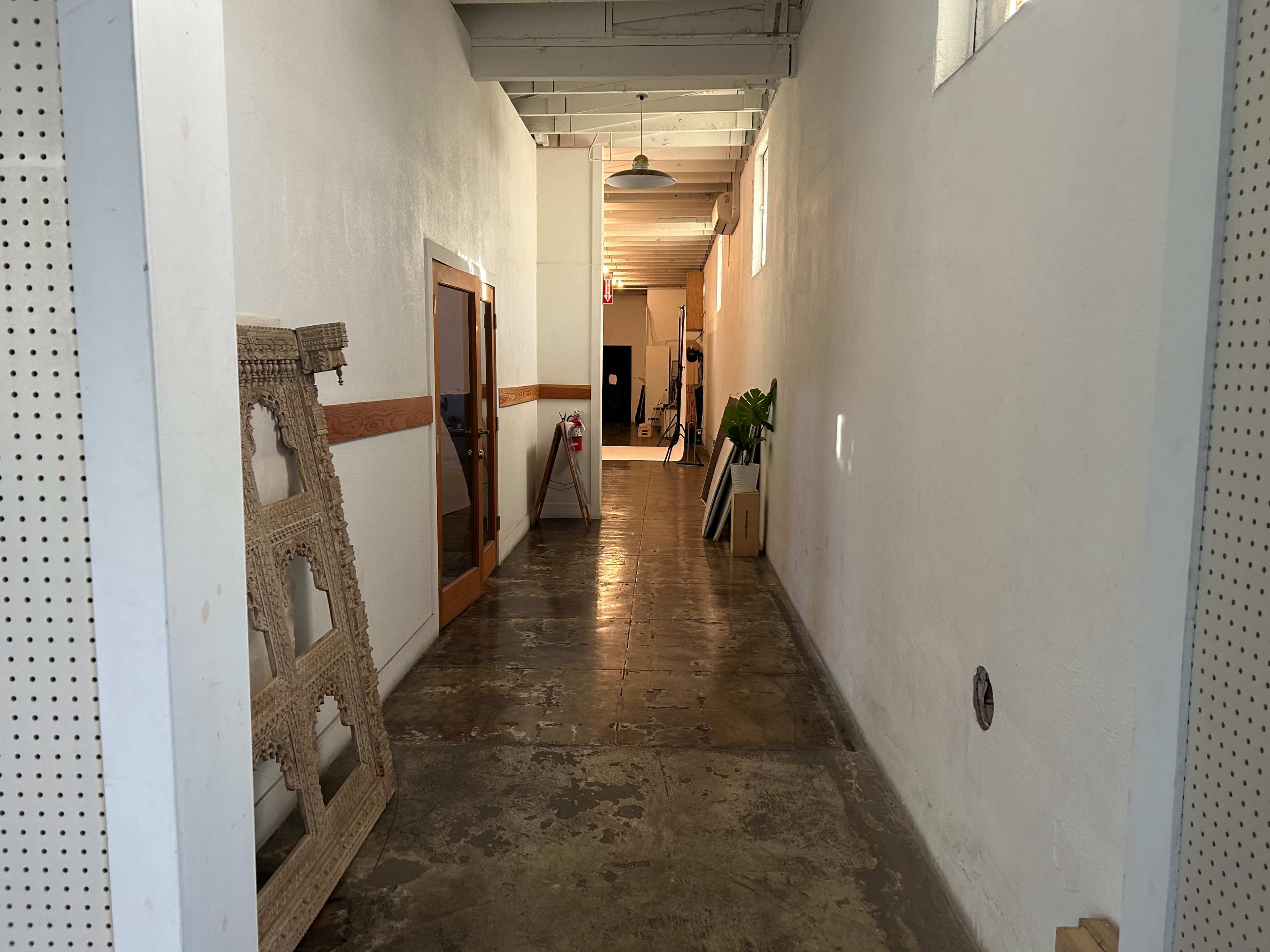 A long corridor with white walls, a polished concrete floor, and various objects, including a large frame and potted plants, lining the sides.