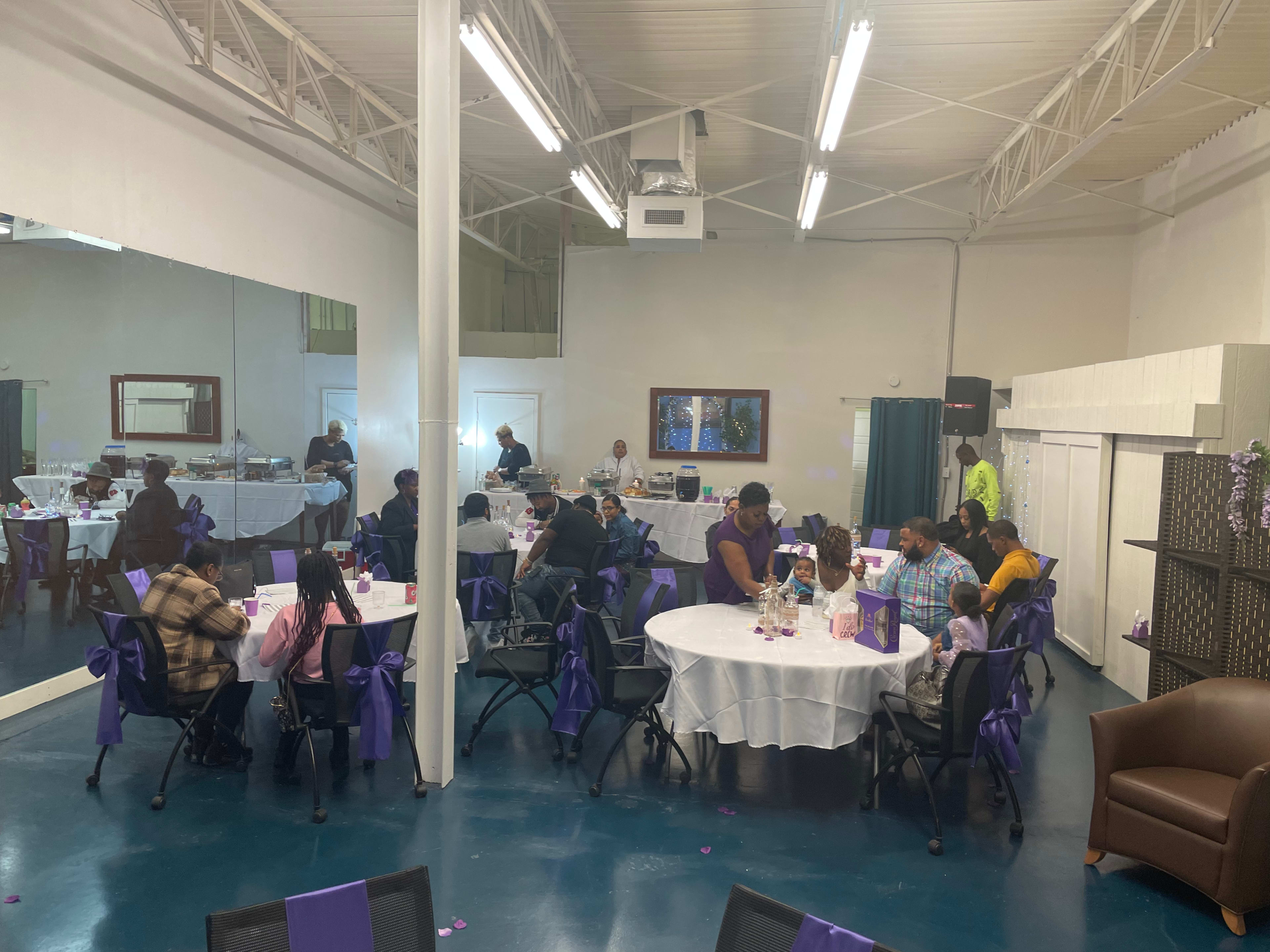 A group of people is seated at tables in a decorated event space with purple accents, enjoying a gathering.