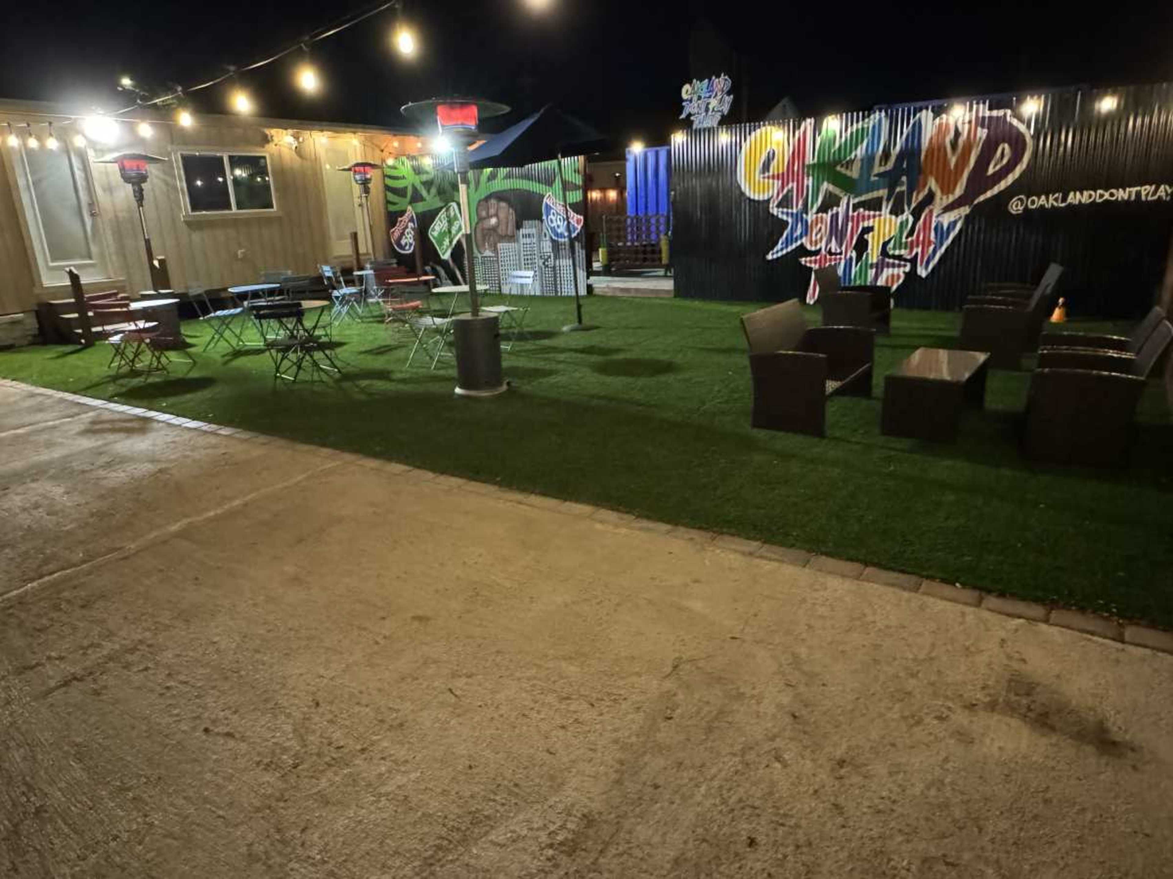 The image shows an outdoor seating area with green grass, scattered tables and chairs, and colorful decorations under string lights at night.