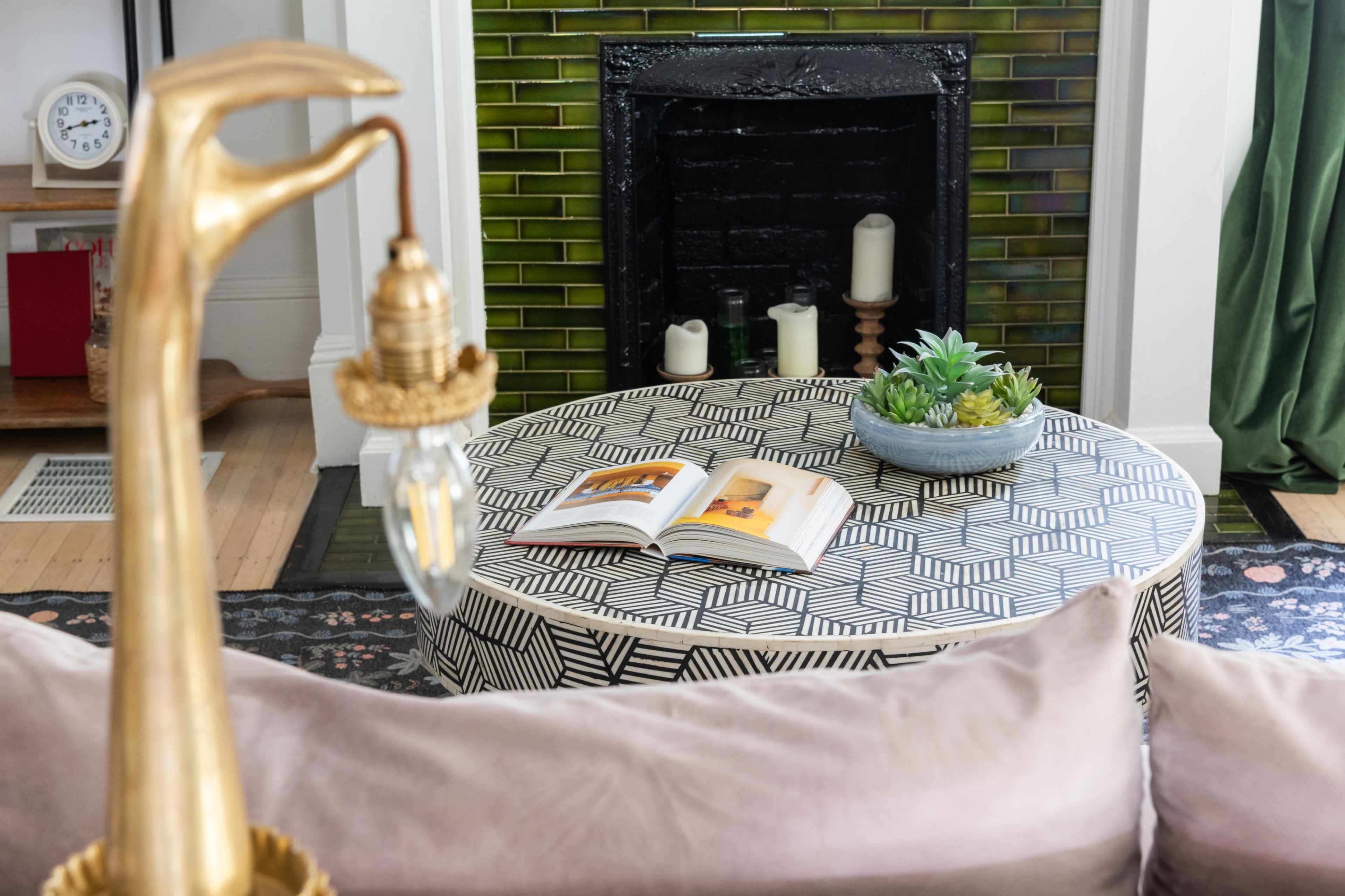 A patterned coffee table with an open book sits in front of a black fireplace surrounded by green tiles and candles.