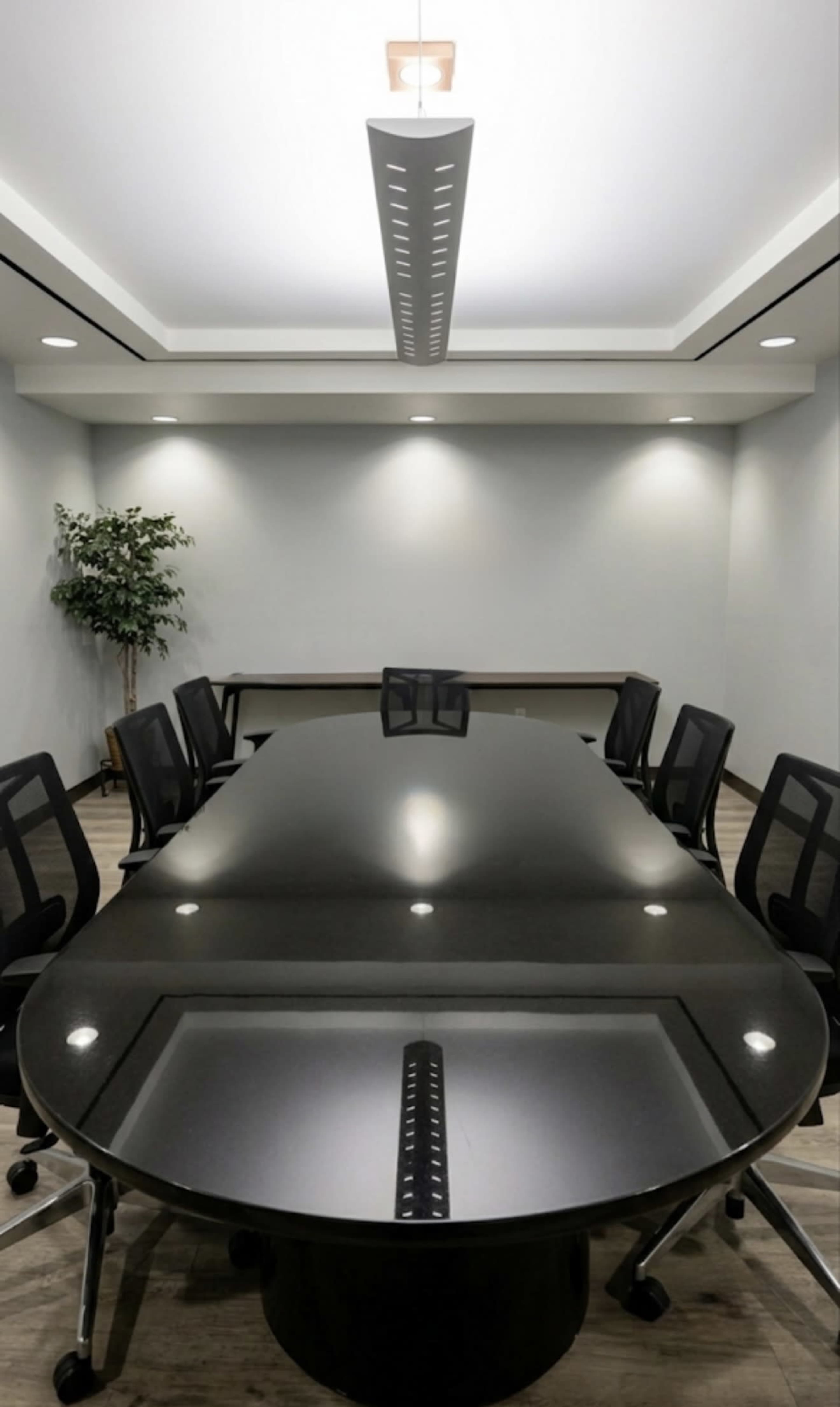 A long, oval conference table surrounded by black rolling chairs is set in a well-lit meeting room with gray walls and a small potted plant in the corner.