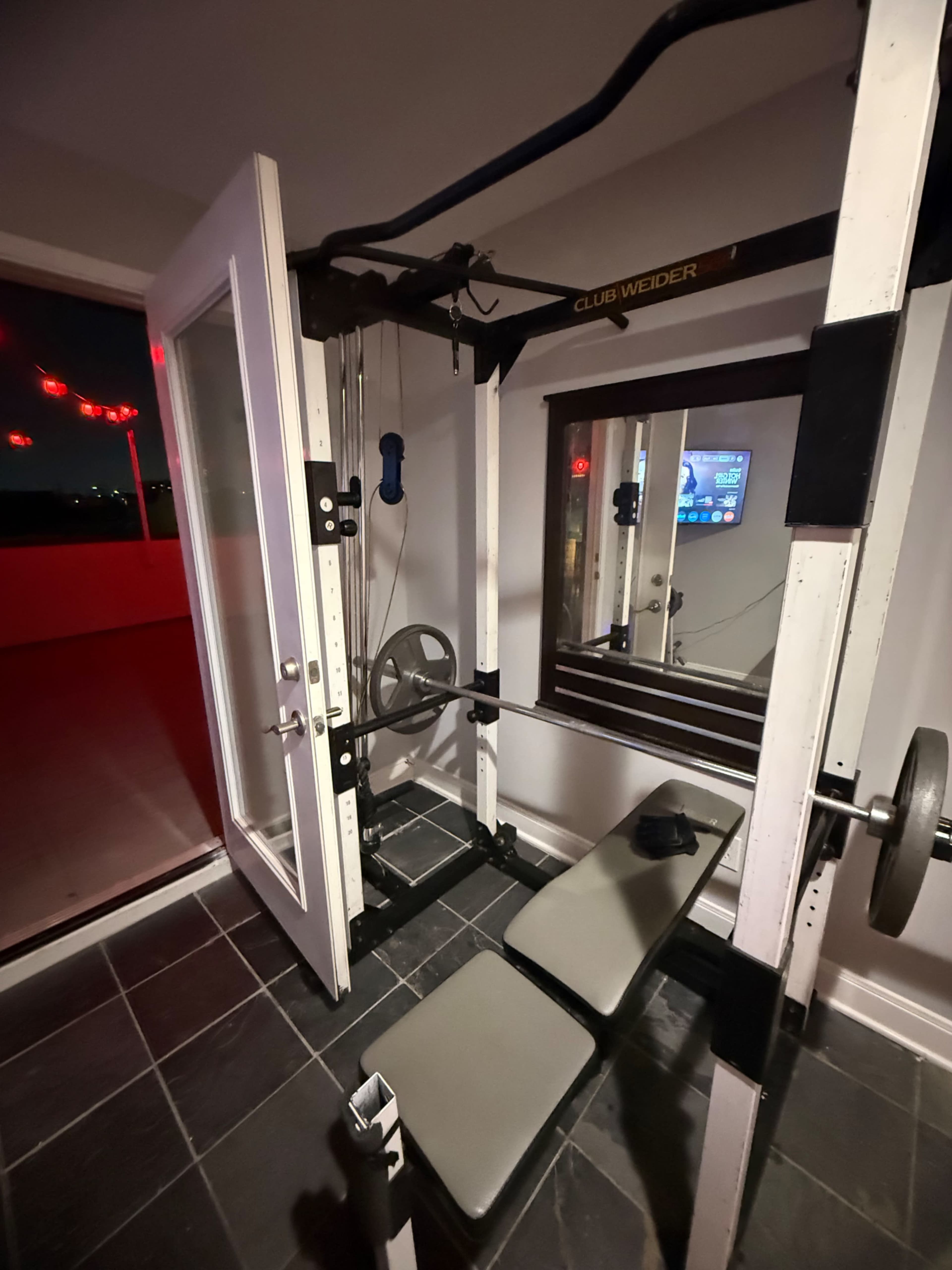 The image shows a weightlifting machine with a bench and barbell setup positioned near a glass door leading outside, reflected in a nearby mirror.