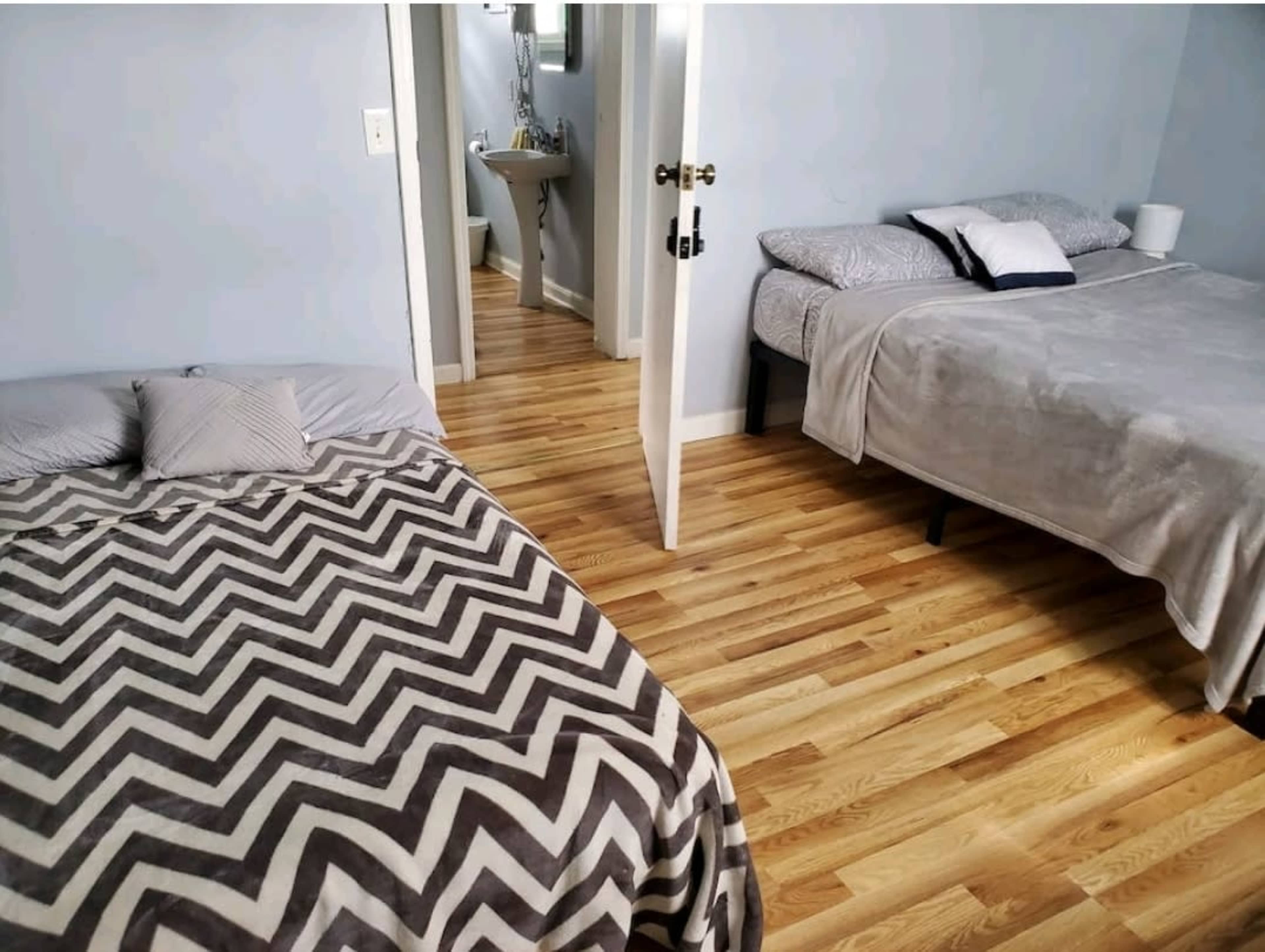 A light blue bedroom with two beds on hardwood floors and a doorway leading to a bathroom in the background.