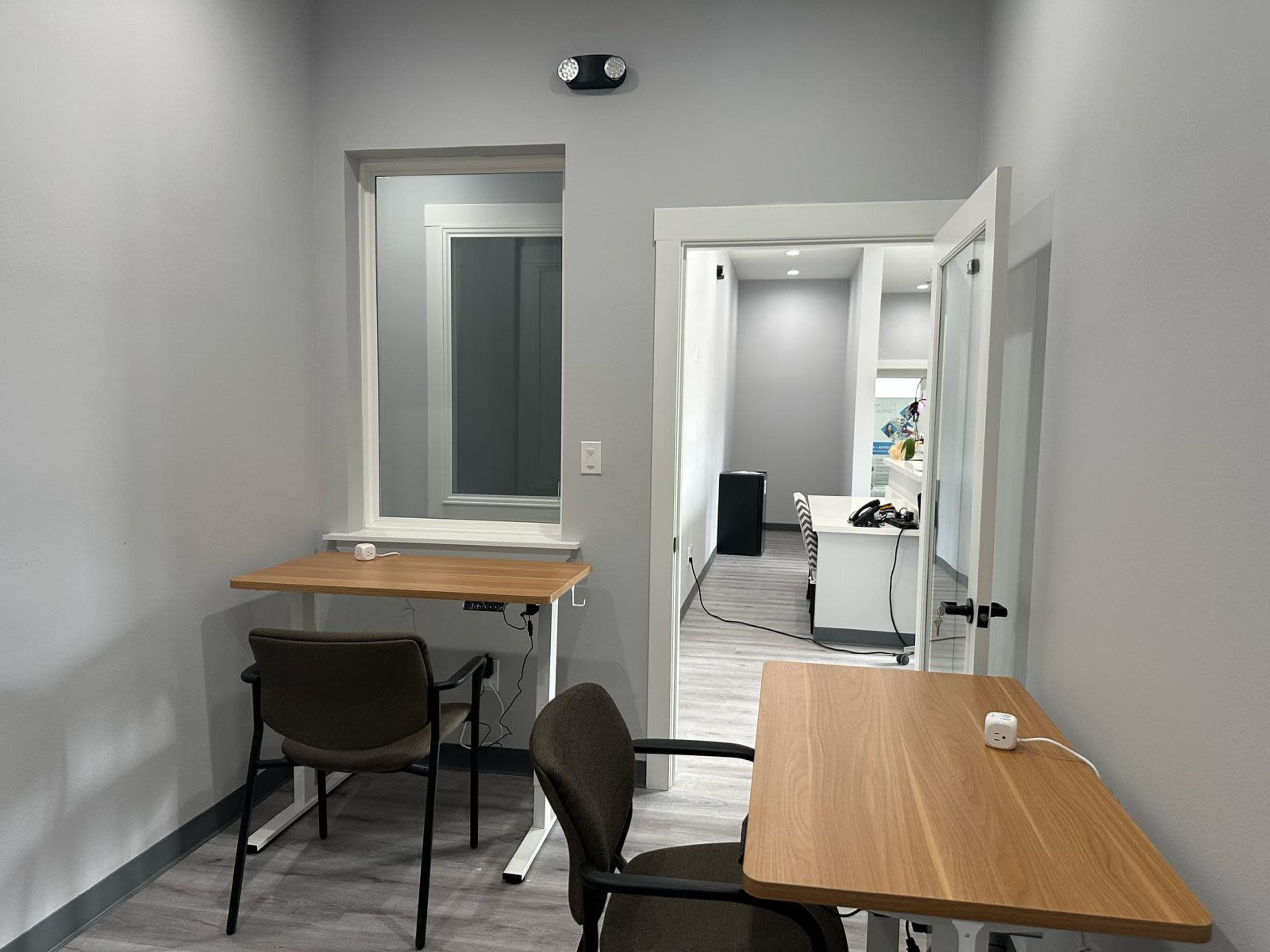 The image shows a small office space with two wooden desks and chairs, one side featuring a door leading to another room.