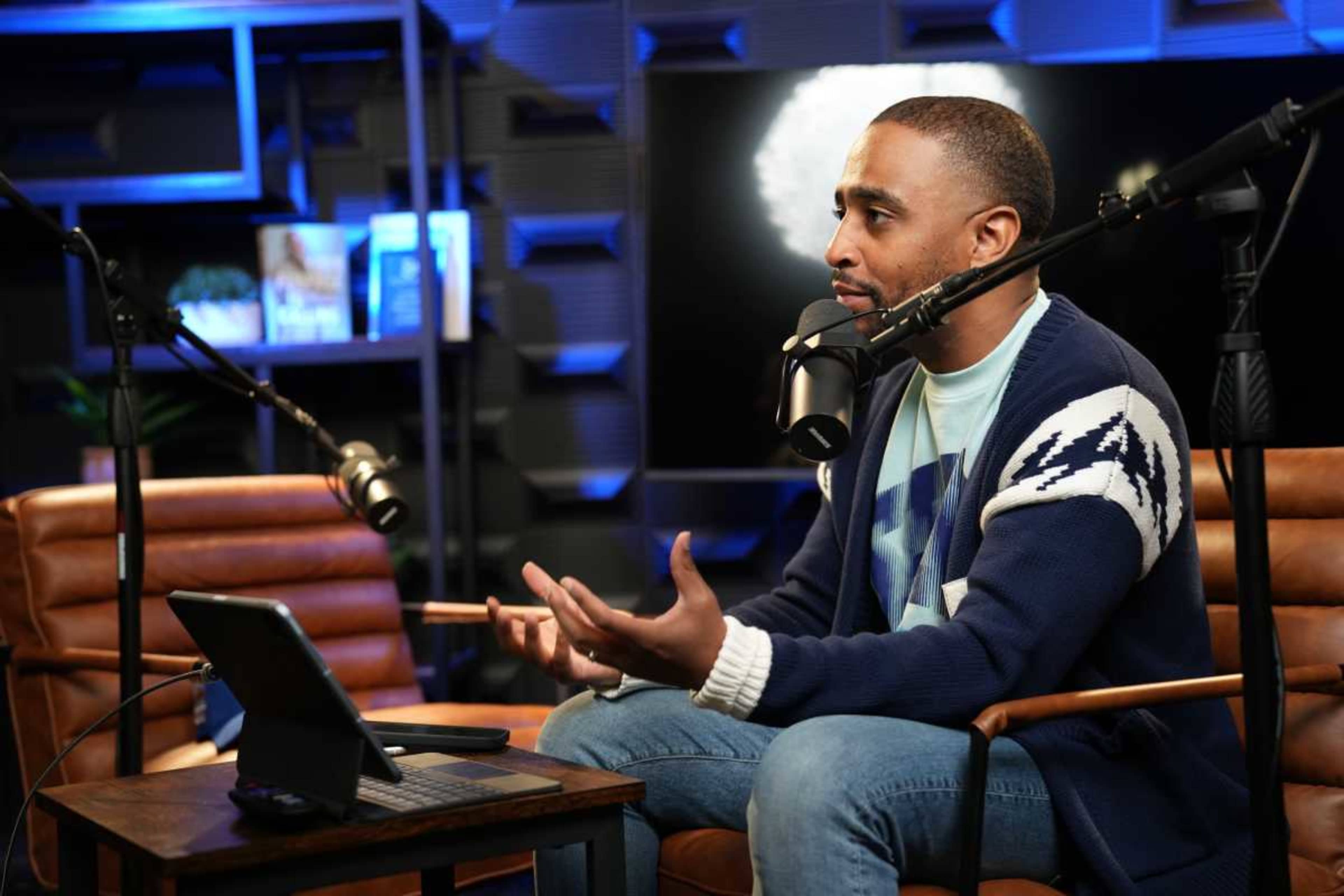 A man is sitting in a studio setting, engaging in conversation while microphone equipment is positioned around him and a tablet is on a table nearby.