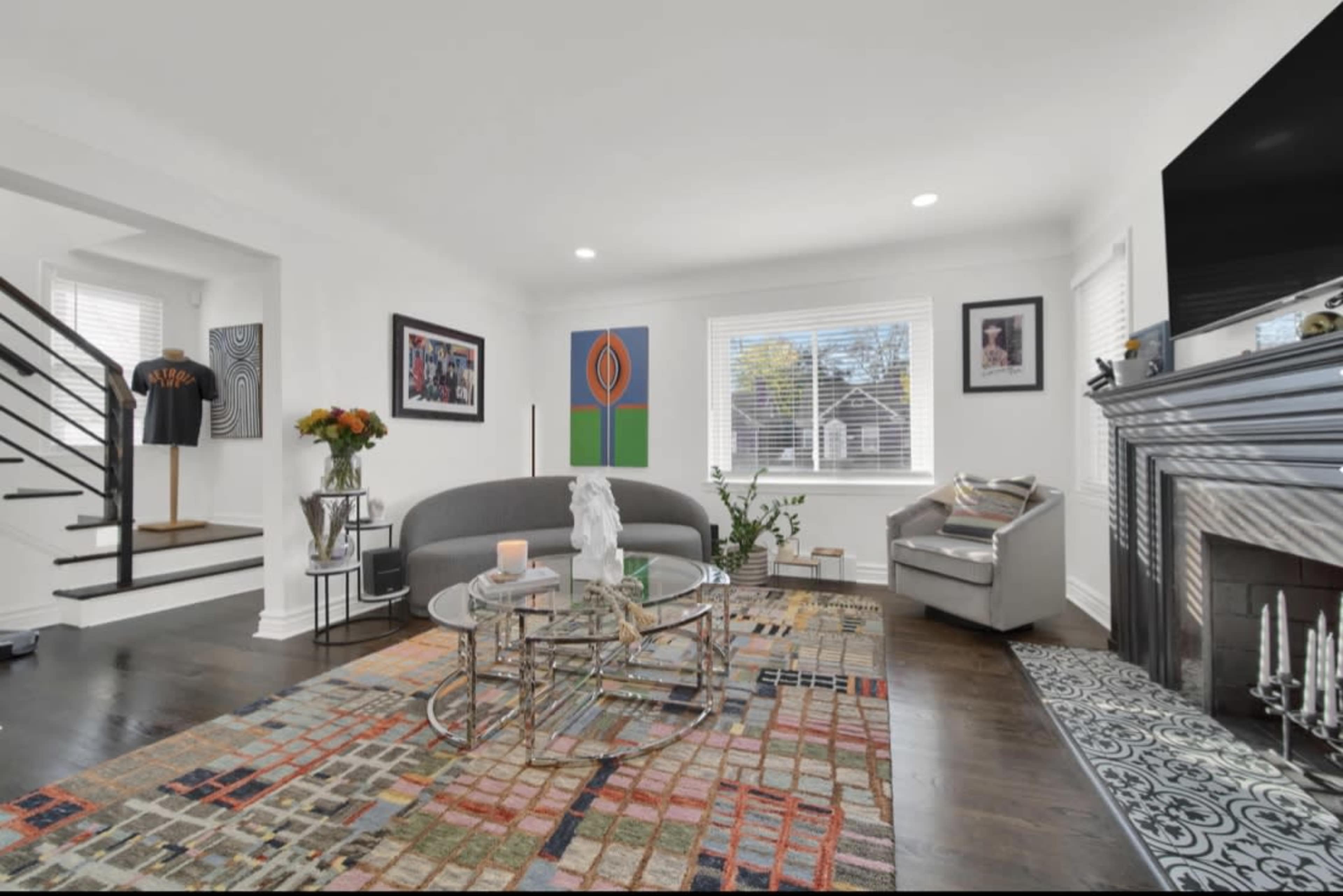 A modern living room features a gray sofa, glass coffee table, and colorful area rug, with artworks on the walls and natural light coming through the windows.
