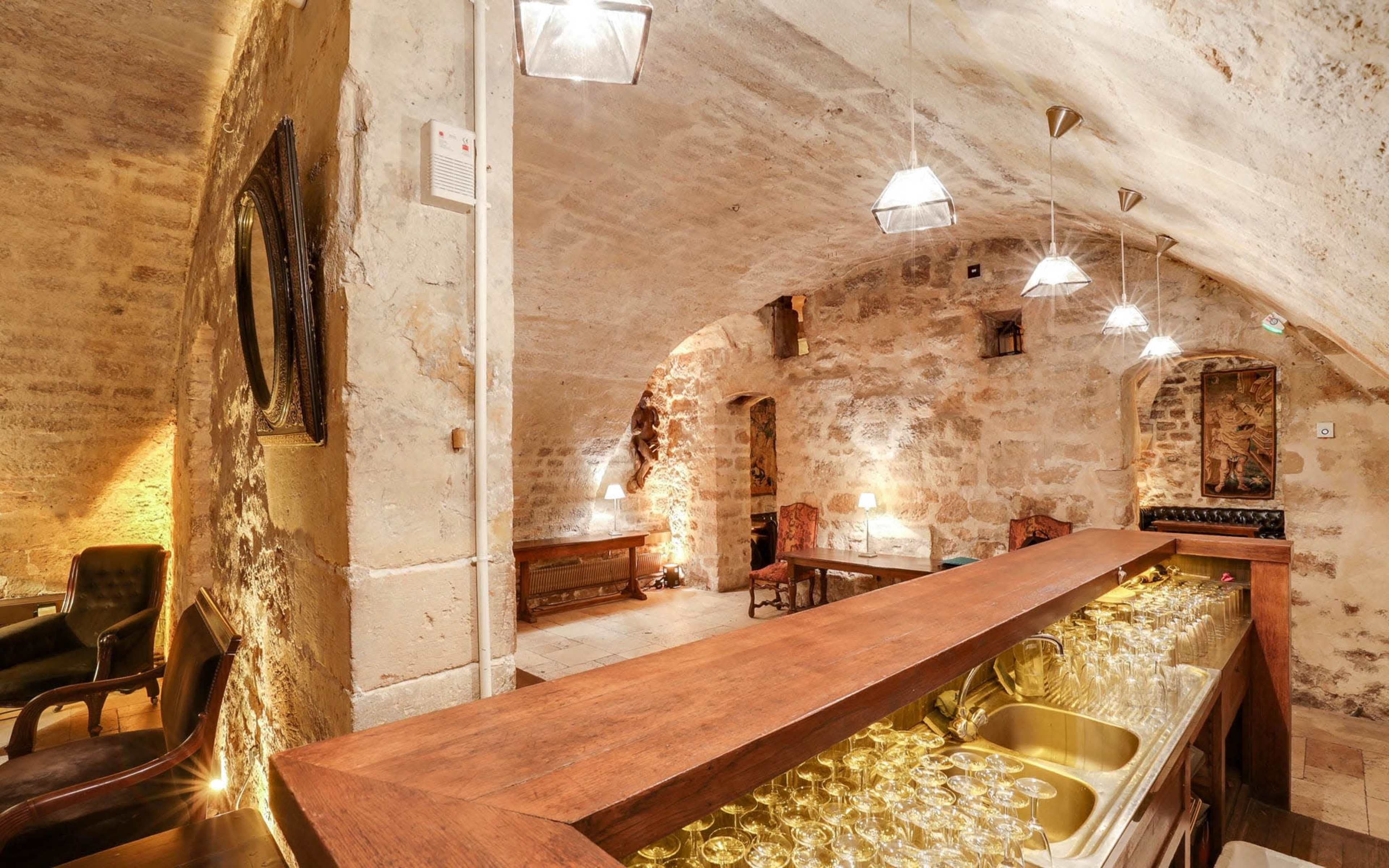 A rustic wine cellar with exposed stone walls, a wooden bar displaying glasses, and low lighting from pendant fixtures.