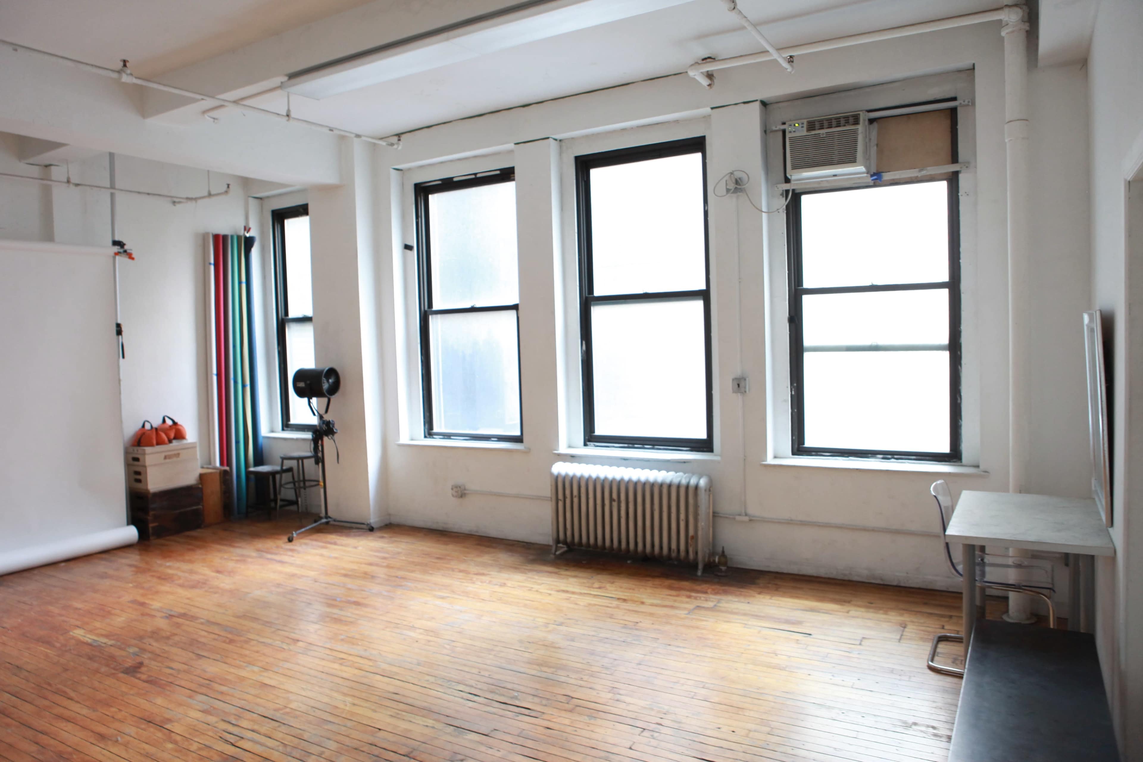 The image shows an empty room with wooden flooring, three large windows, and minimal furnishings, including a radiator and a small table.