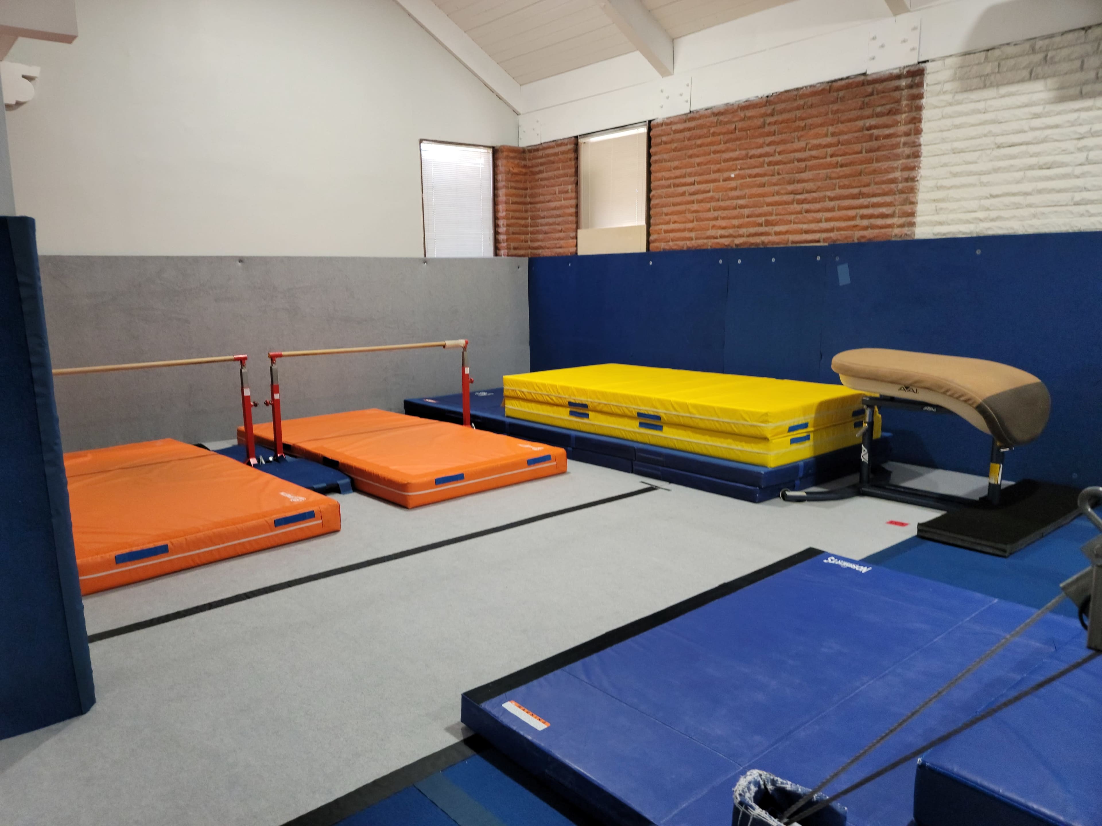 A gymnasium training area featuring orange and blue mats, uneven bars, a vaulting table, and a springboard arranged on a gray floor.