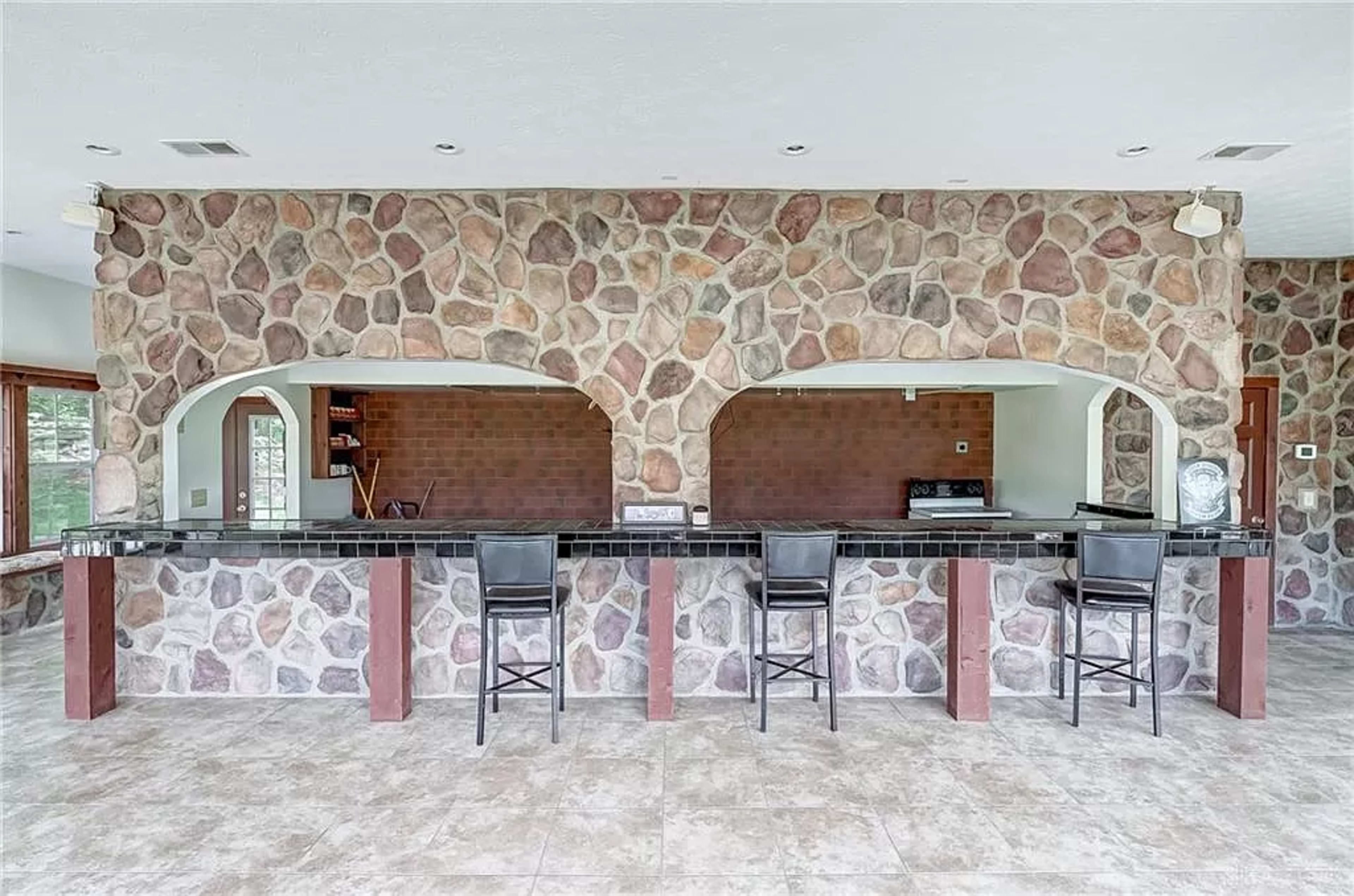 A spacious bar area featuring a stone wall with arched openings and four black barstools positioned along a dark countertop.