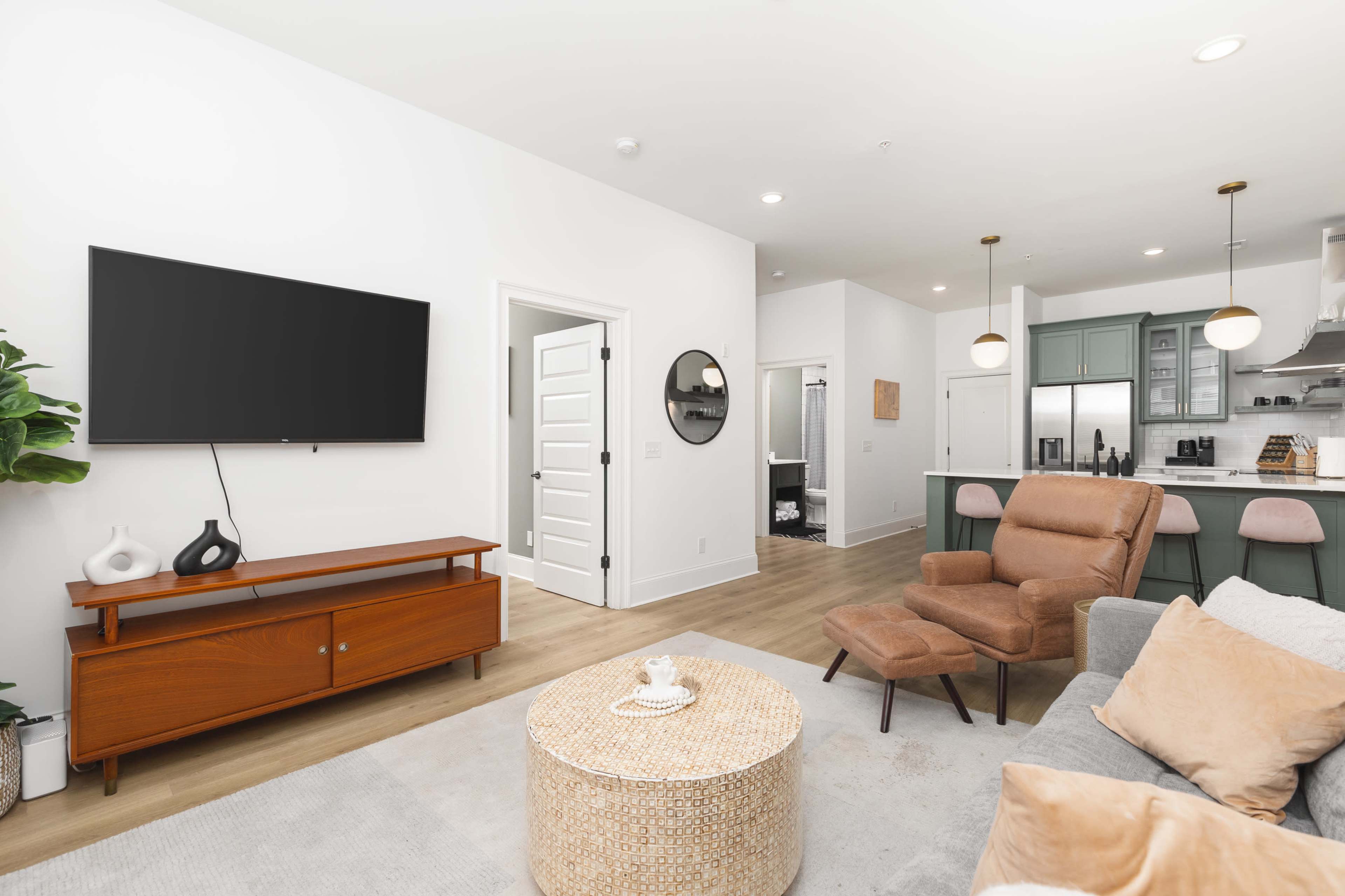 A modern living room features a large TV mounted on a white wall, a wooden media console, an armchair, a round woven coffee table, and an open kitchen in the background.