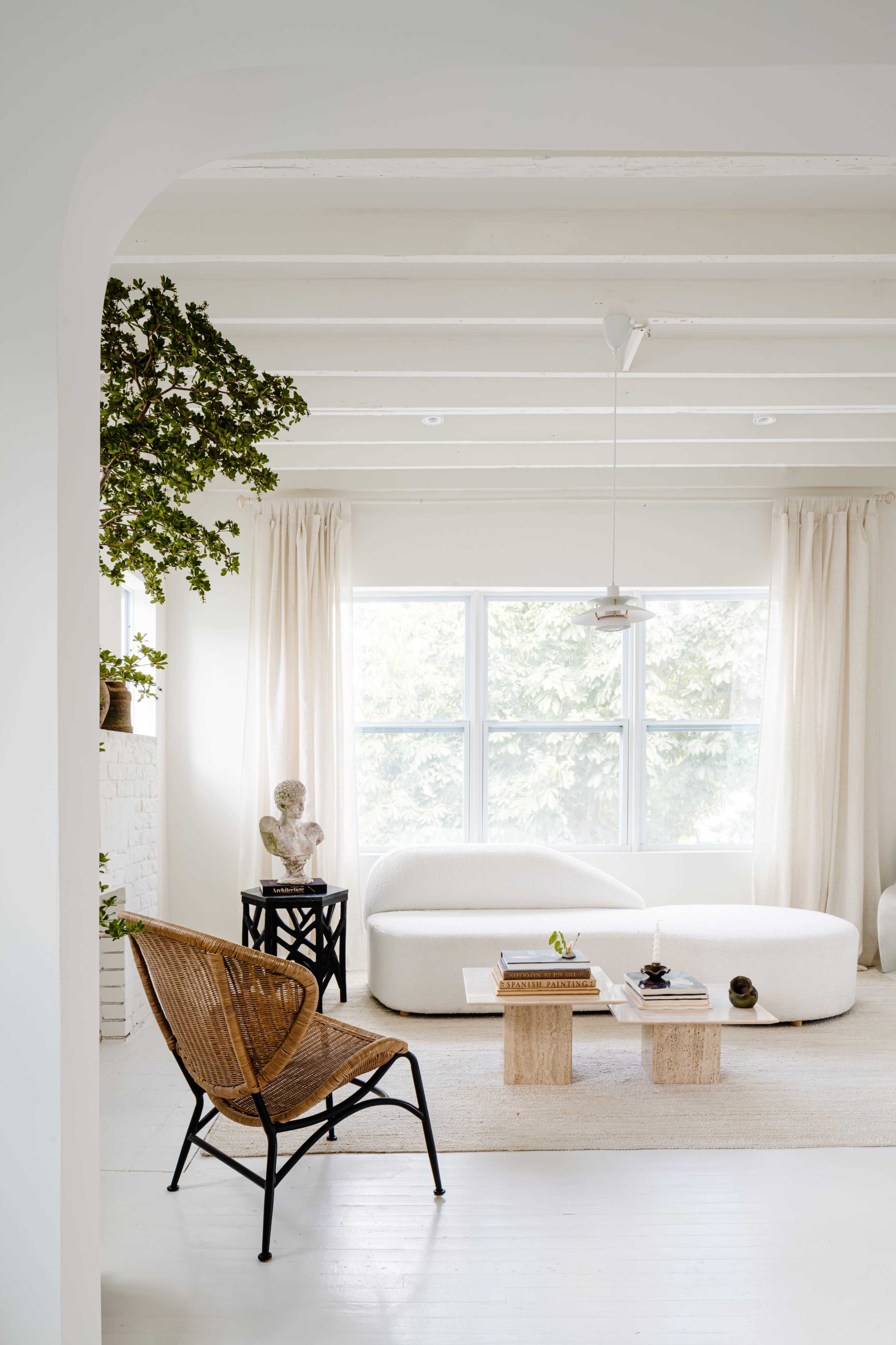 A bright, minimalist living room features a white sofa, a wooden coffee table, a rattan chair, and large windows with sheer curtains.
