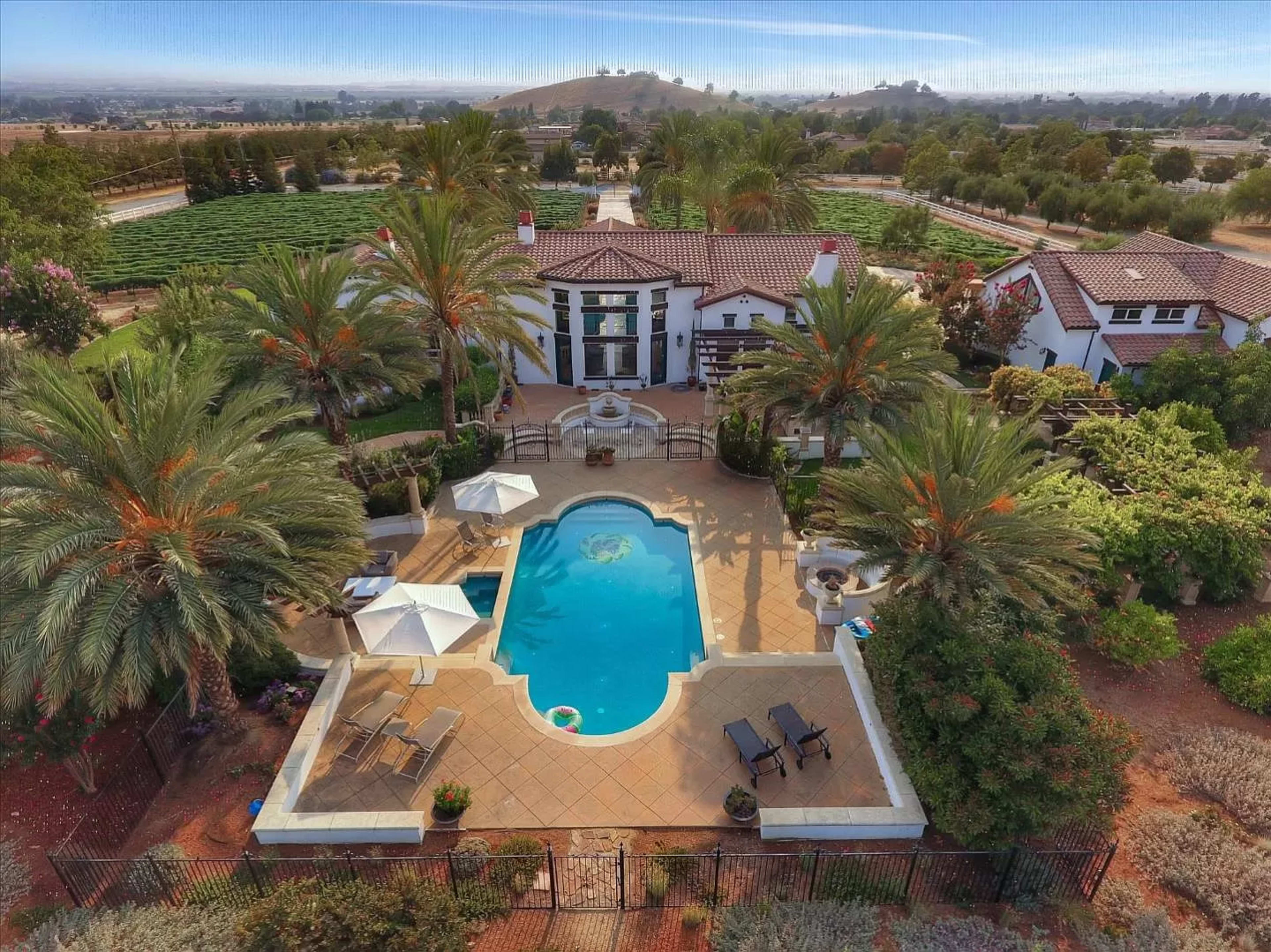 A landscaped property with a swimming pool, surrounded by palm trees, lounge chairs, and a patio, set against a backdrop of rolling hills.