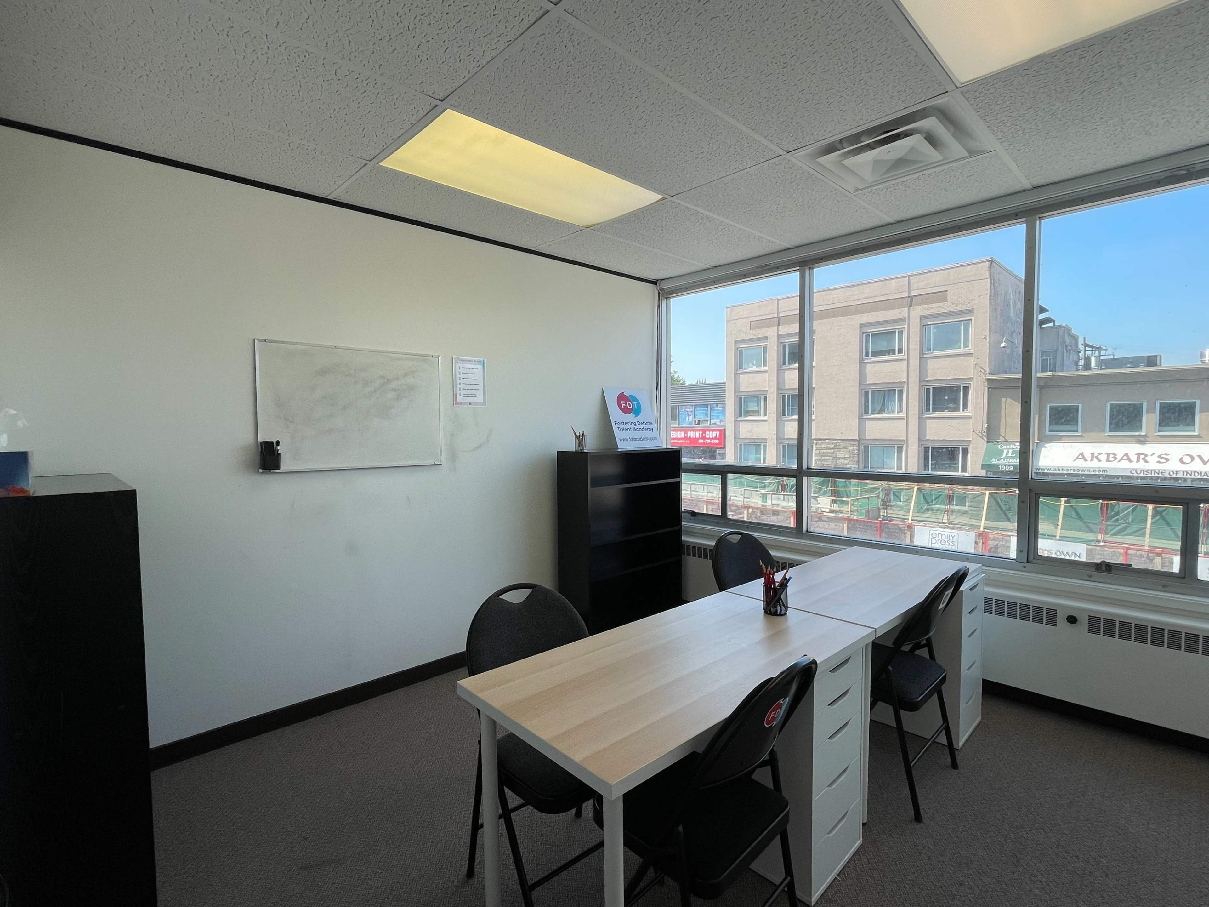 A small office space features a table with chairs, a whiteboard on one wall, and a large window overlooking a city view.
