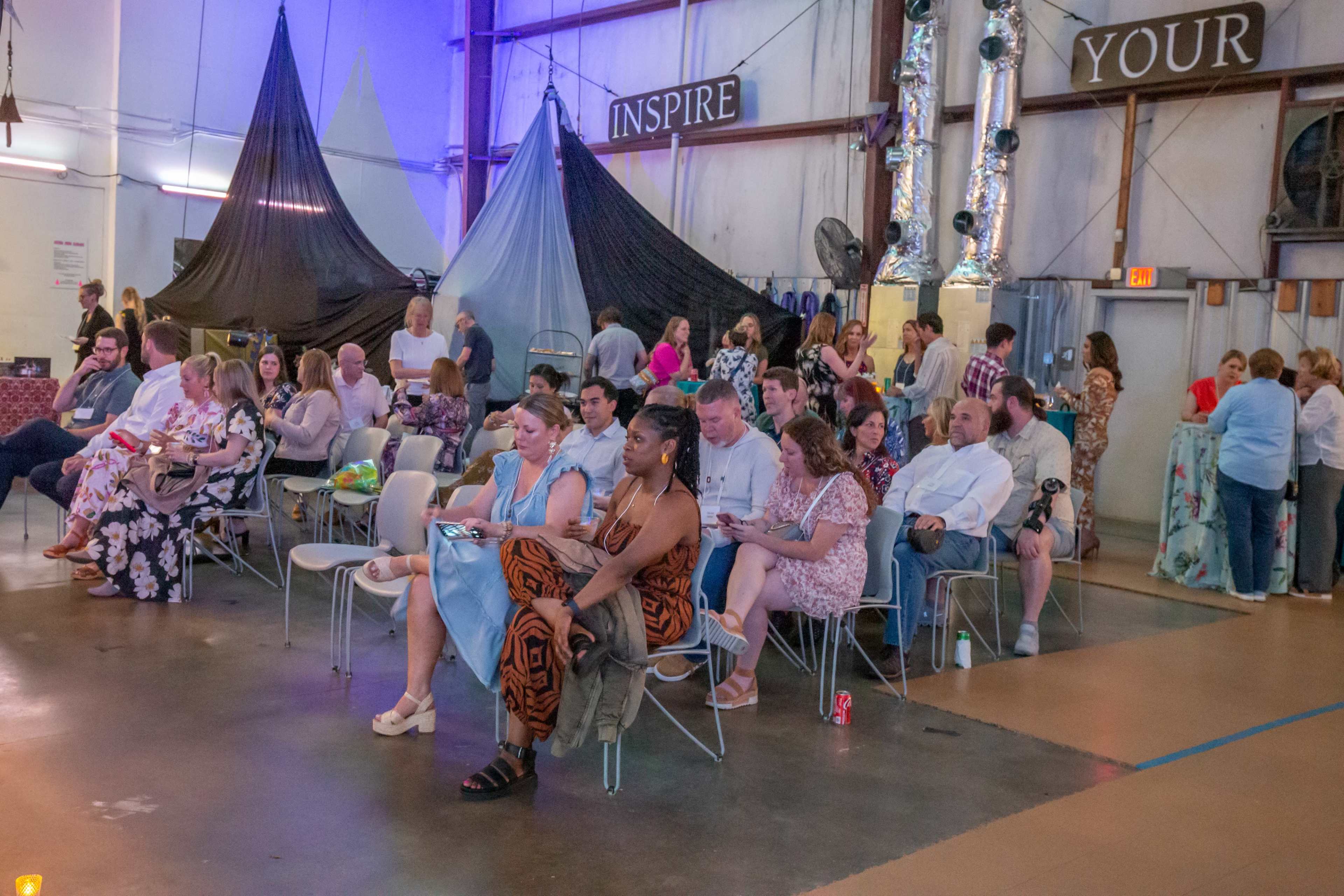 A varied group of people sits in a large indoor space, some engaged in conversation, while others look at their phones, with fabric draped above creating distinct sections.