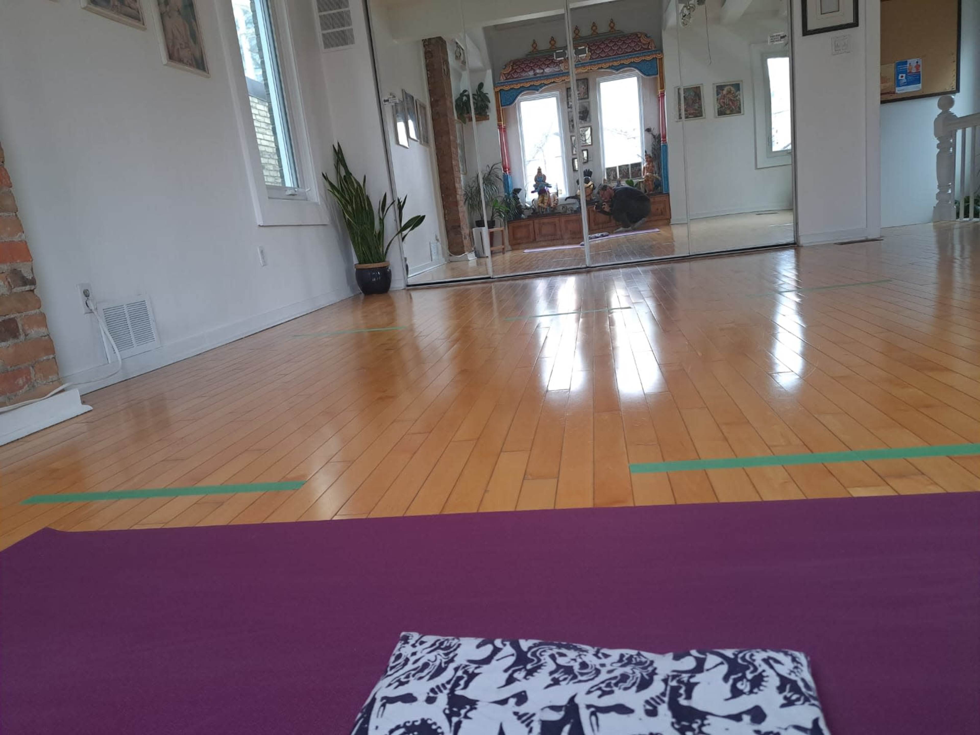 A yoga mat is placed on a polished wooden floor in a spacious room with large mirrors and plants.