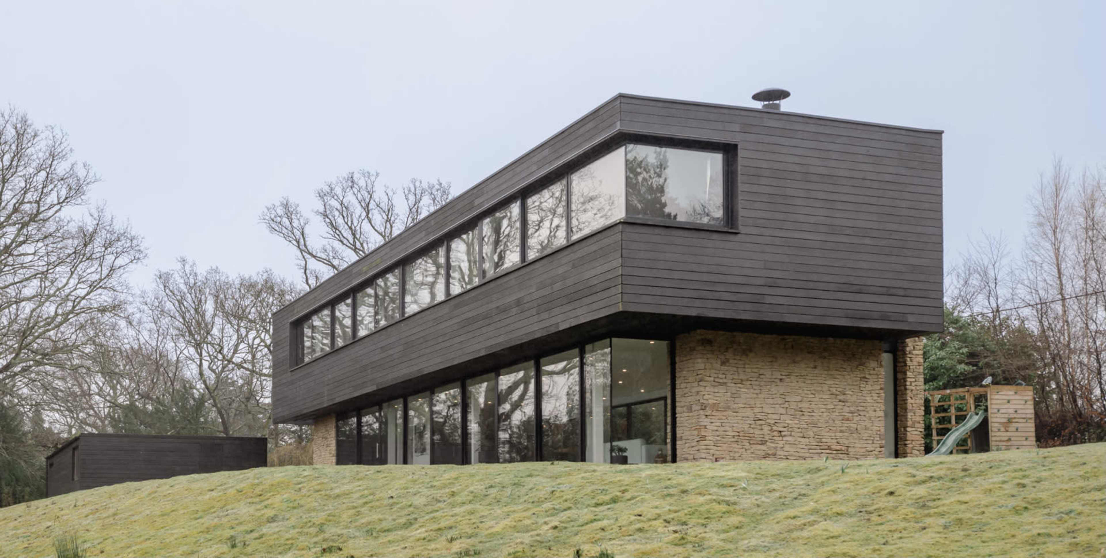 A modern two-story house with a combination of stone and black wood siding is situated on a gently sloped grassy area.