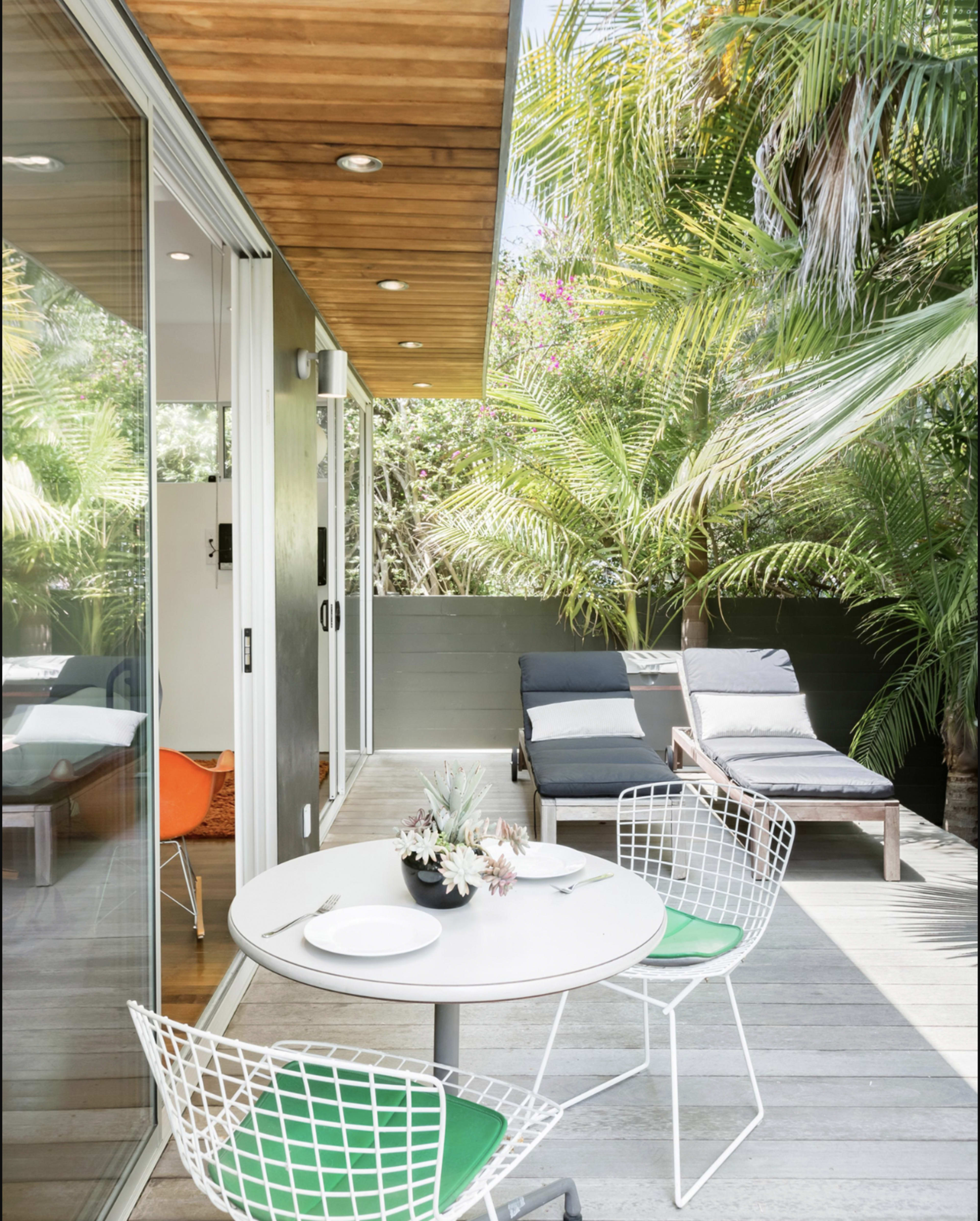 The image shows a modern outdoor patio with a small round table and chairs, surrounded by lush greenery and two lounge chairs.