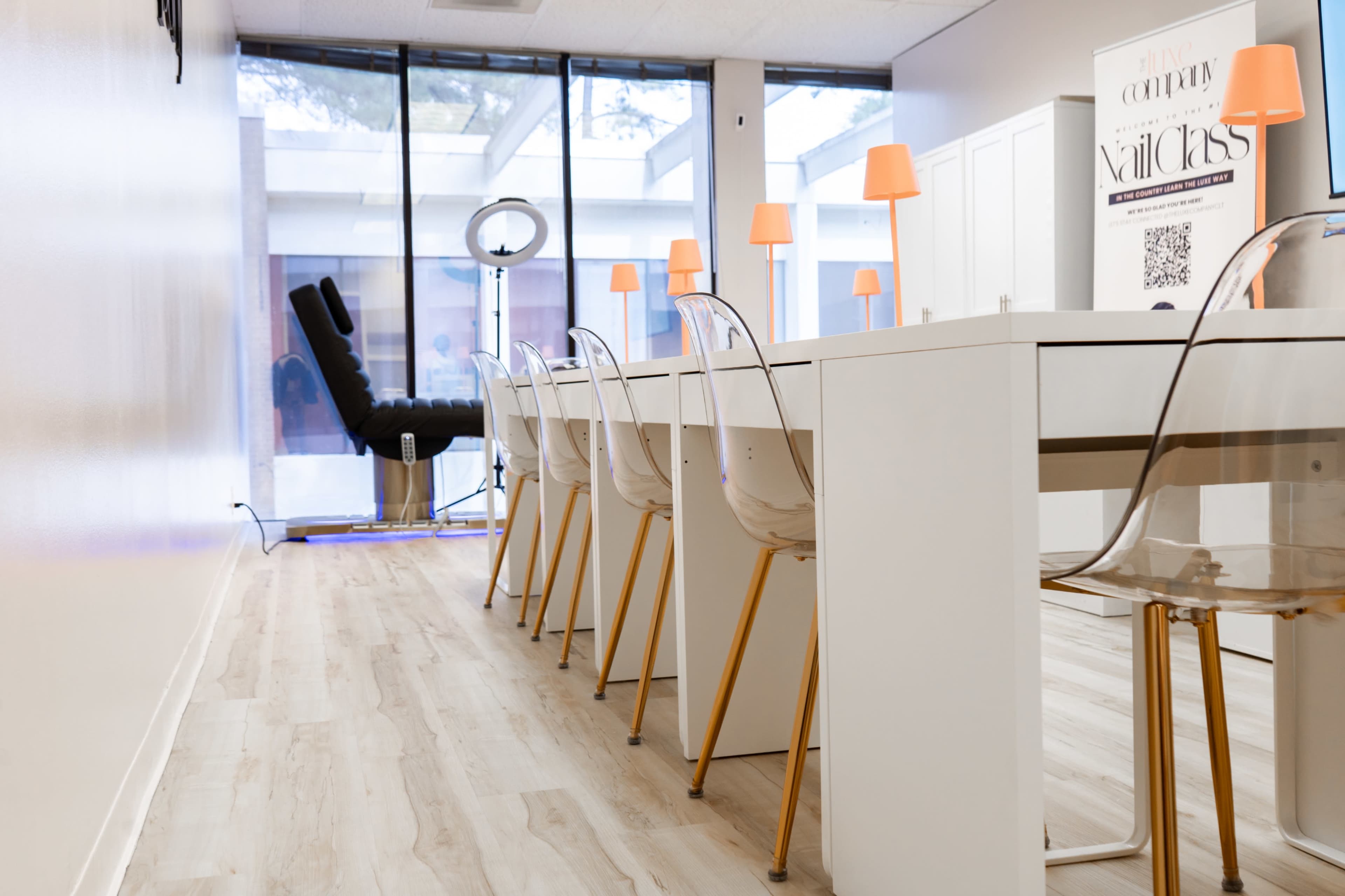 The image shows a modern, minimalistic workspace with a long white table, clear plastic chairs, and orange lamps along the sides, leading to a well-lit area with large windows.