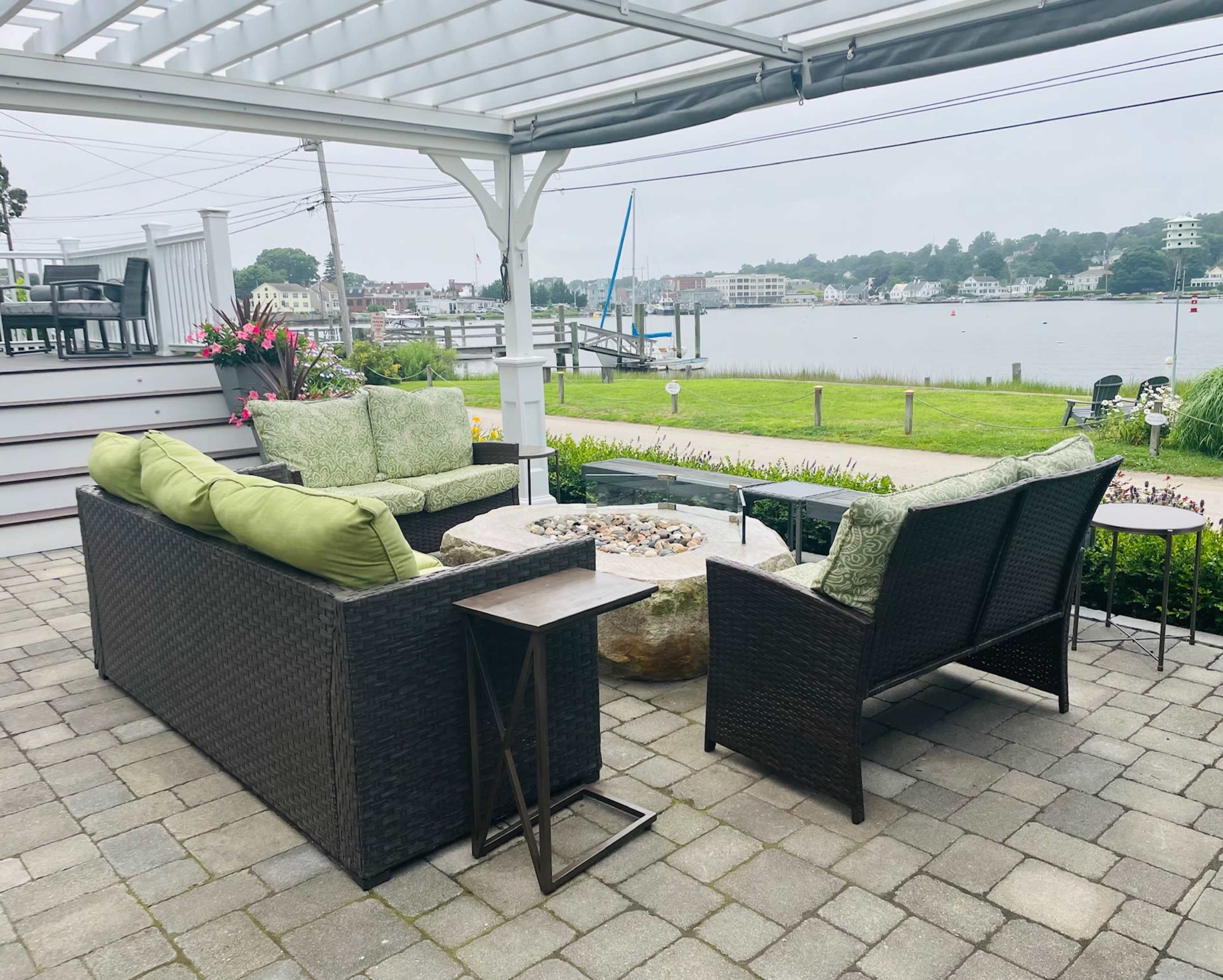 A seating area with green cushions overlooks a waterfront, featuring a stone fire pit surrounded by paver stones.