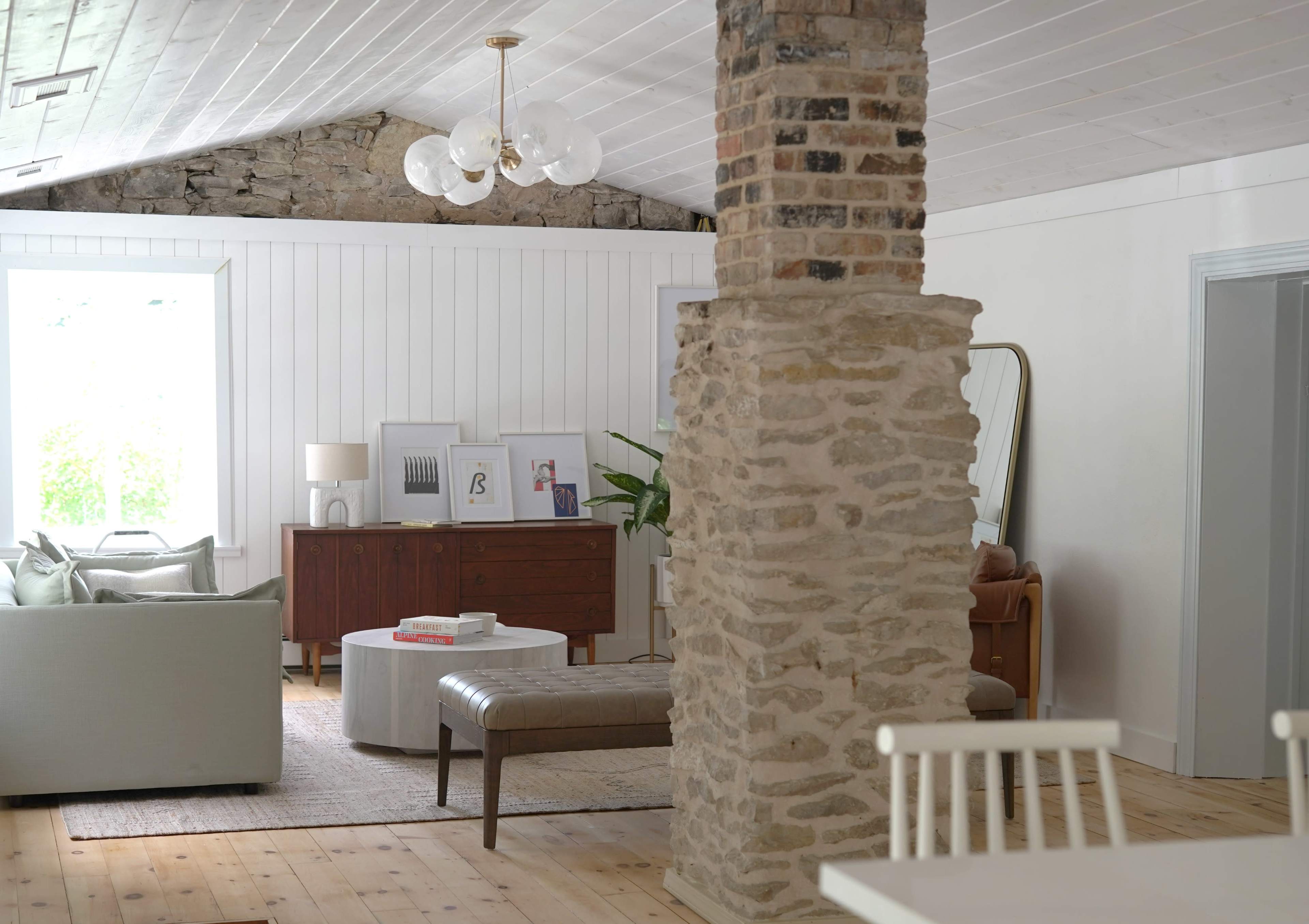 The image depicts a spacious living room featuring a stone column, a light-colored sofa, a round coffee table, and a mix of modern and vintage furnishings.