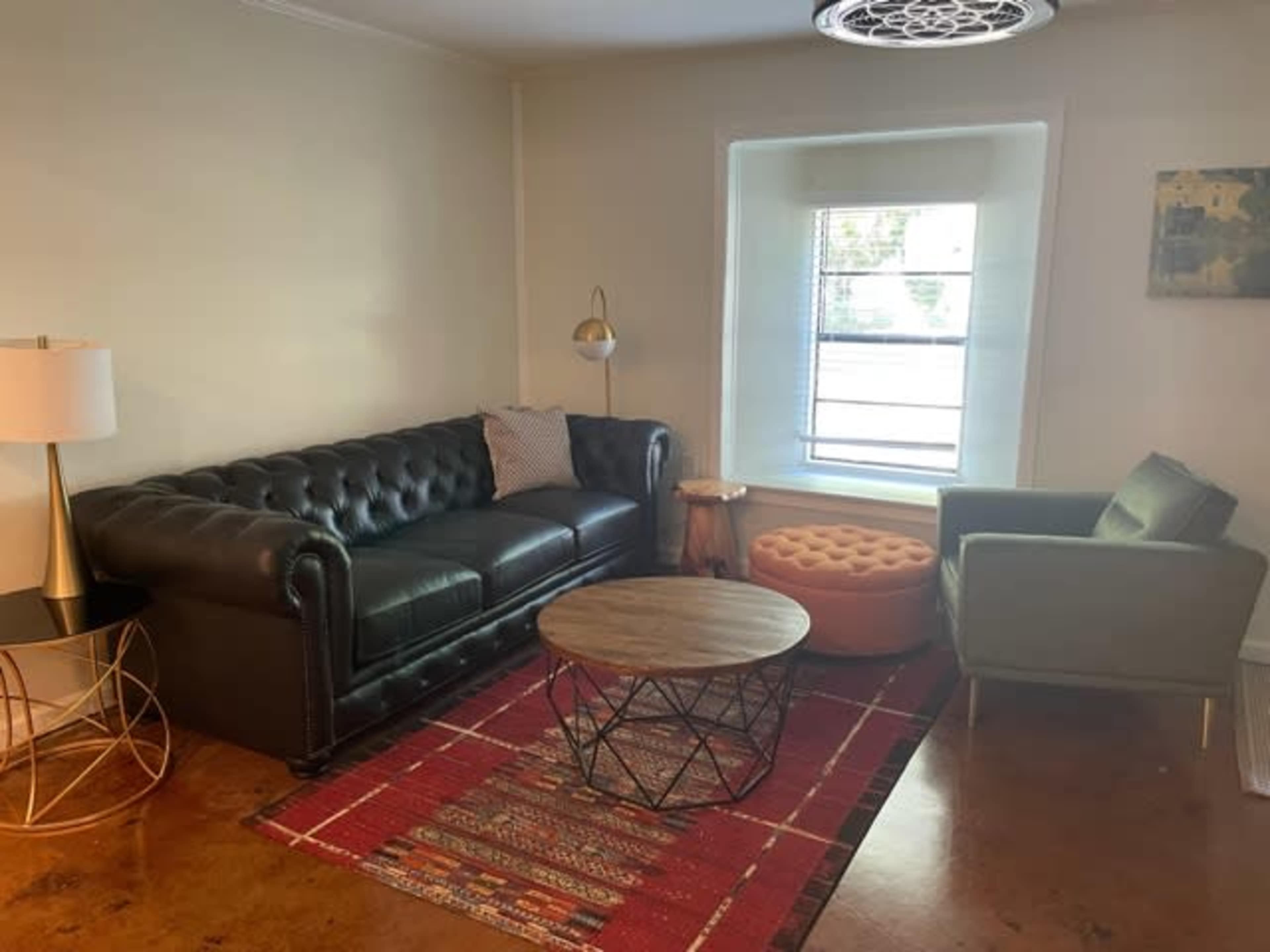 A living room with a black leather tufted sofa, a round wooden coffee table, an orange ottoman, a green armchair, and a light fixture overhead.
