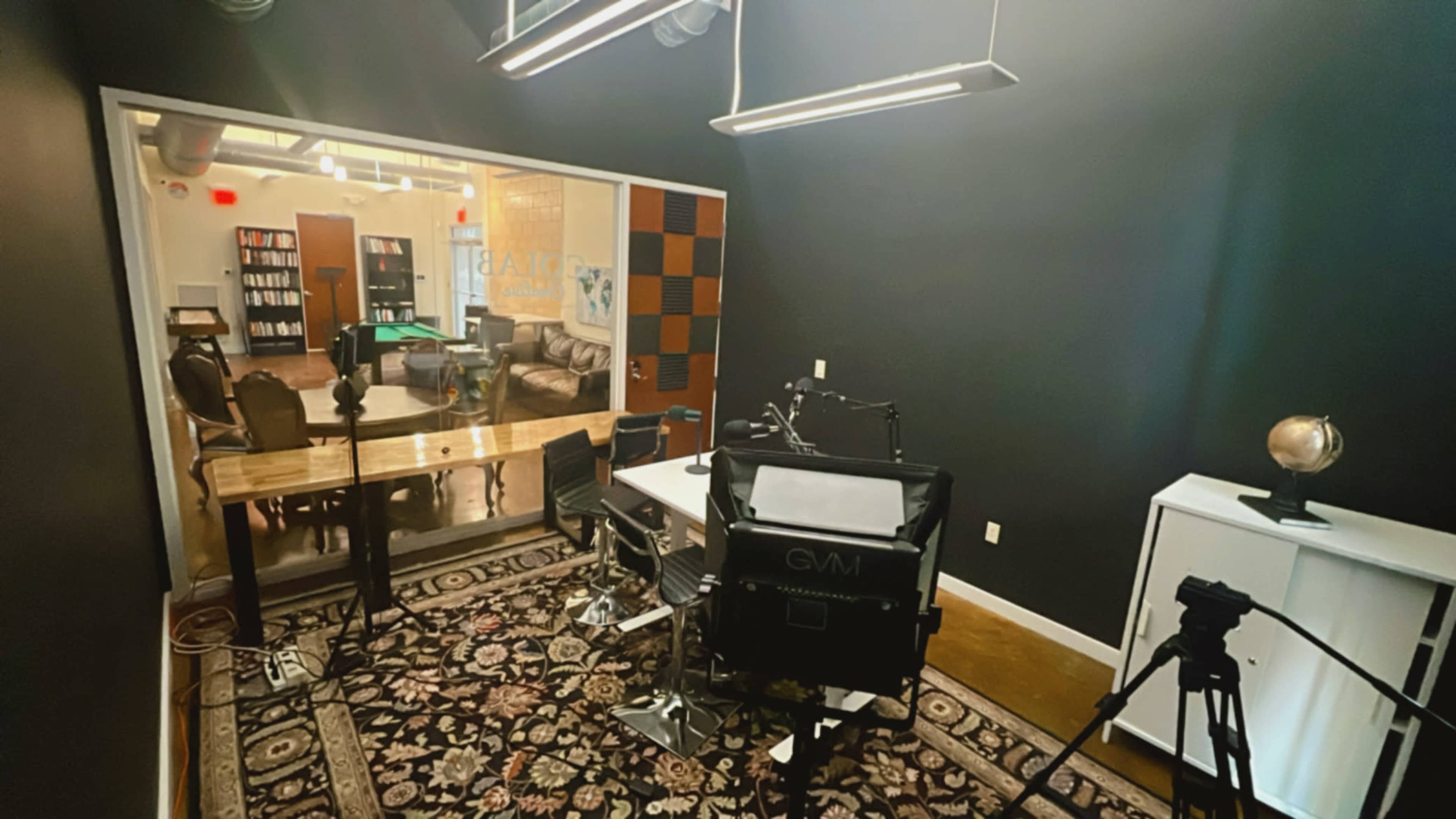 A modern recording studio with a large desk, chairs, and cameras facing a glass-walled lounge area featuring a pool table and bookshelves.