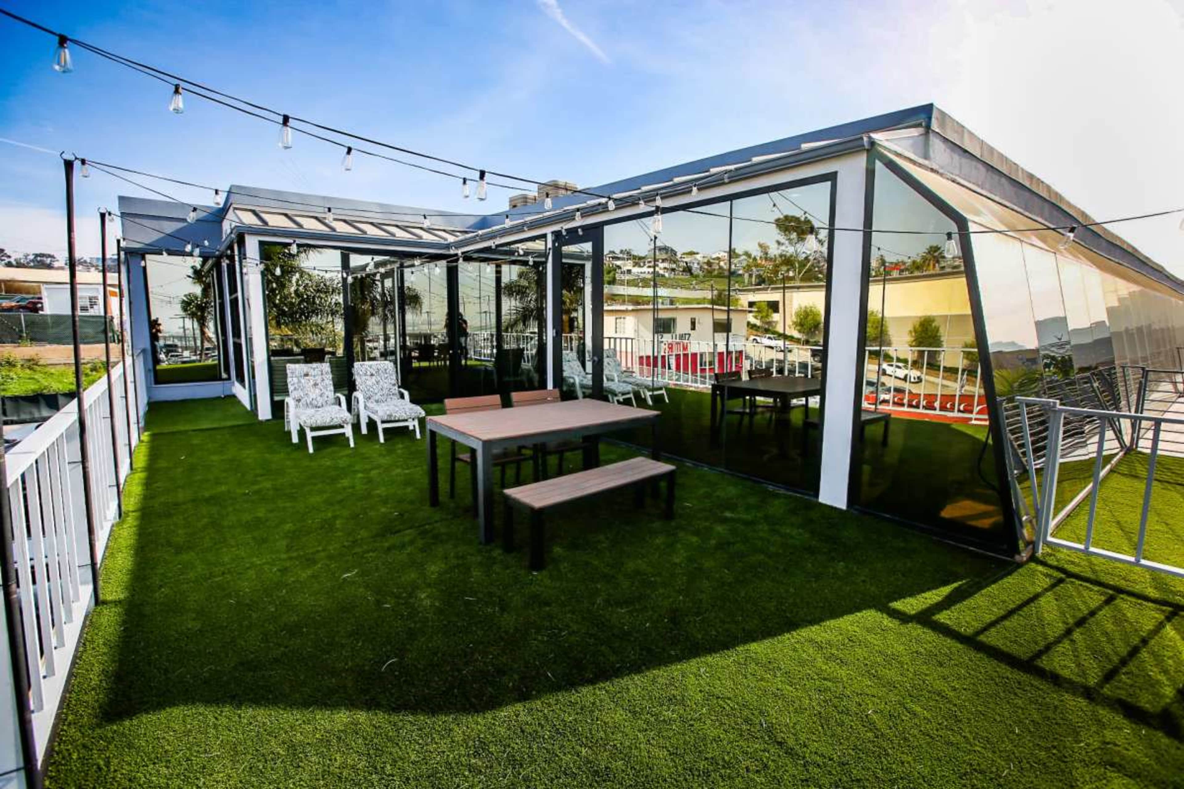The image shows a rooftop terrace with a green artificial lawn, a wooden table with benches, and lounge chairs, surrounded by glass walls and overhead string lights.