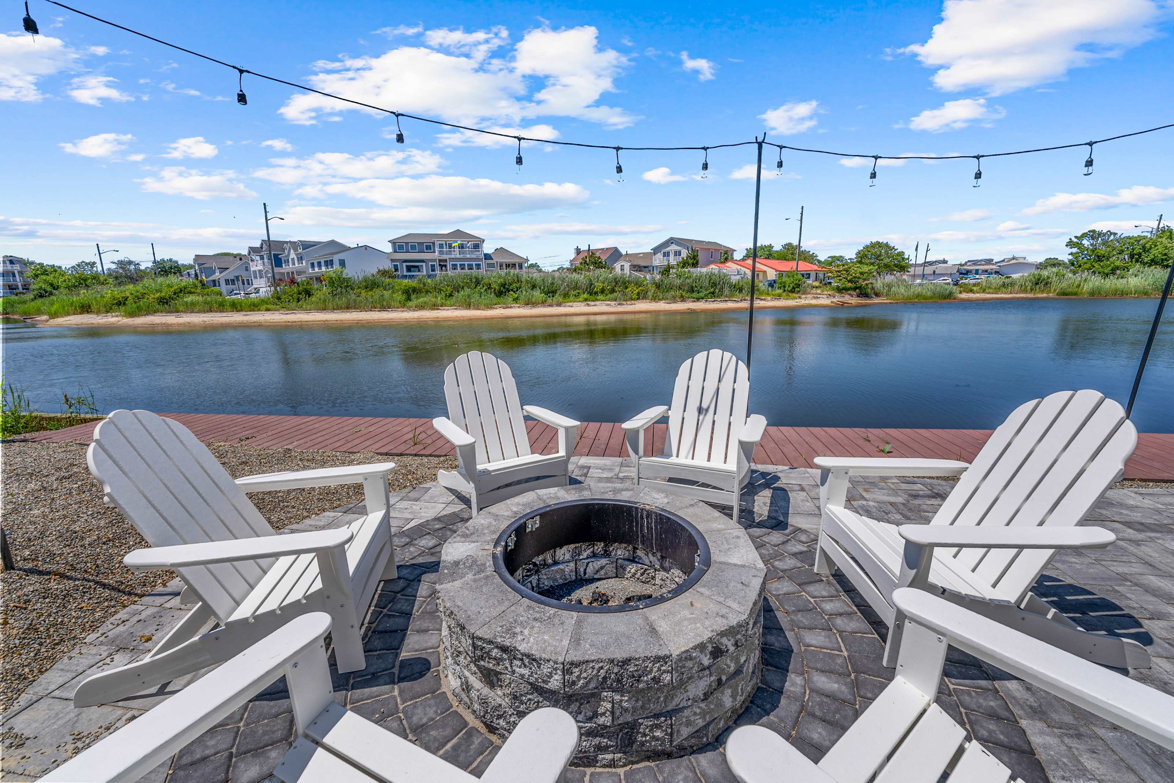 A circle of white Adirondack chairs frames a stone fire pit beside a calm body of water, with houses visible in the background.