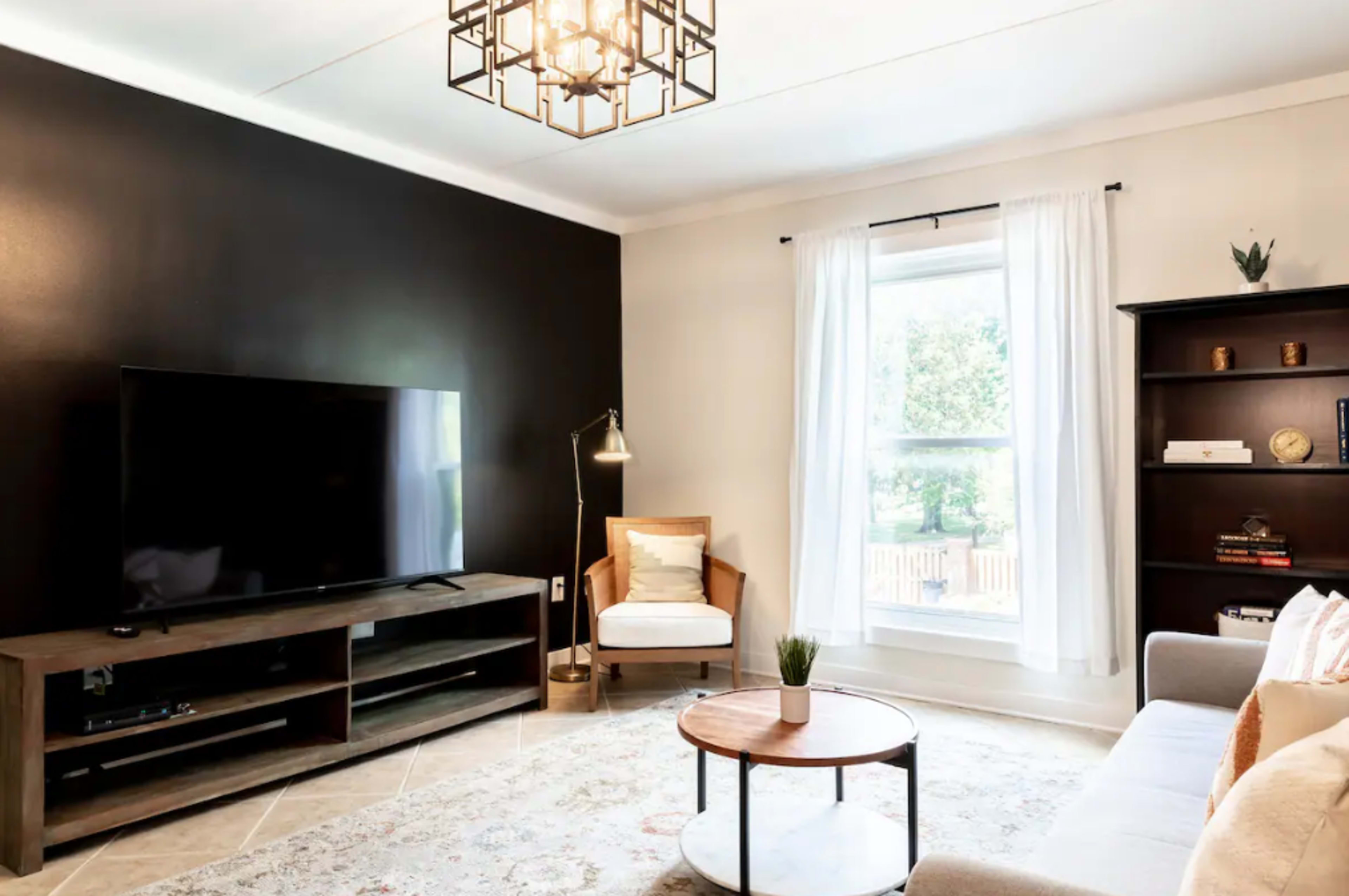 The living room features a television on a wooden stand, a light-colored sofa, a round coffee table, and a large window with sheer curtains.