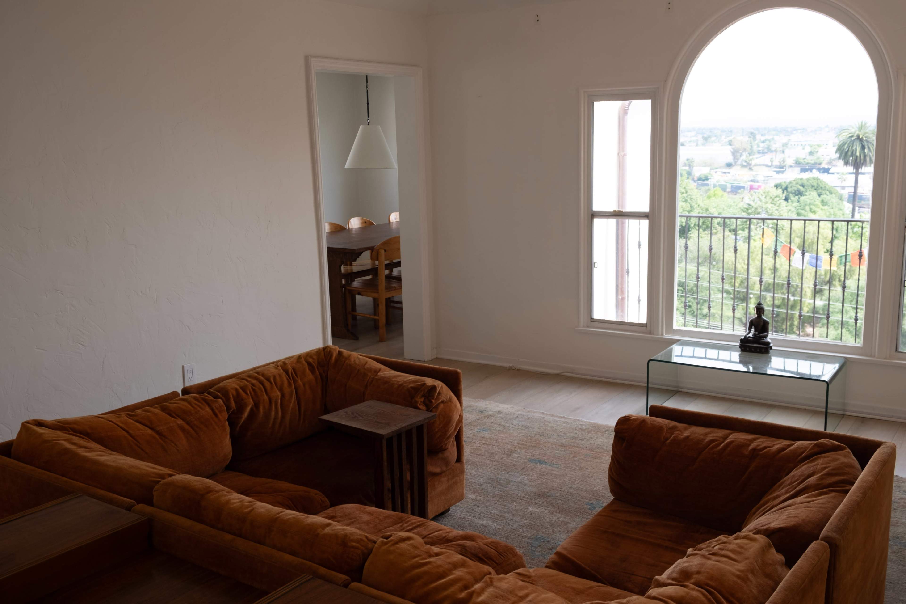The image shows a living room featuring an orange sectional couch, a small wooden side table, and a glass display case, with large windows offering a view of the outside.