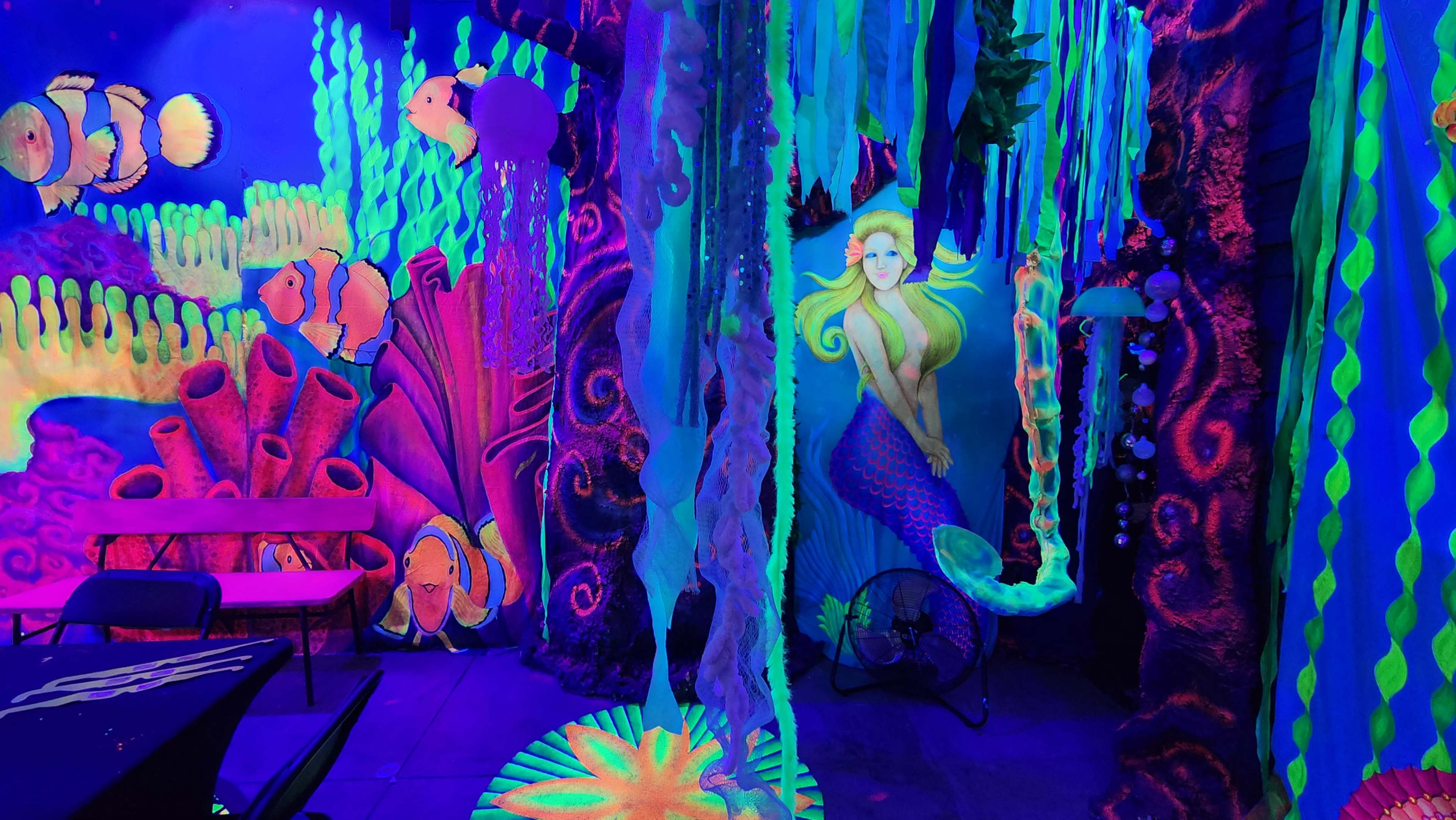 The scene features a vibrant, underwater-themed room illuminated with black light, showcasing colorful murals of fish, sea creatures, and a mermaid among flowing seaweed.