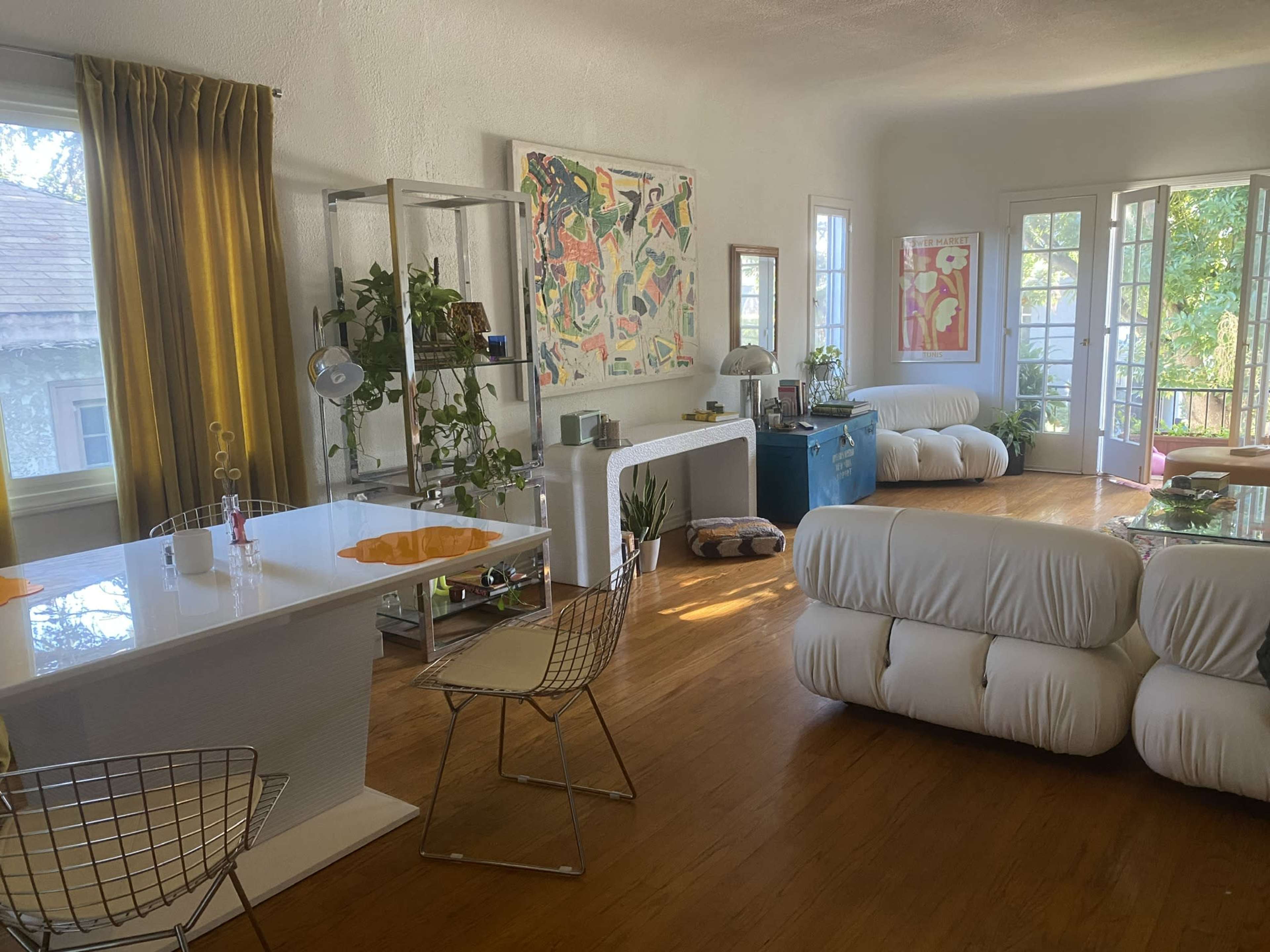 A bright living room with a dining area featuring a white table, metal chairs, and large windows allowing natural light, along with various colorful artworks and furniture pieces.