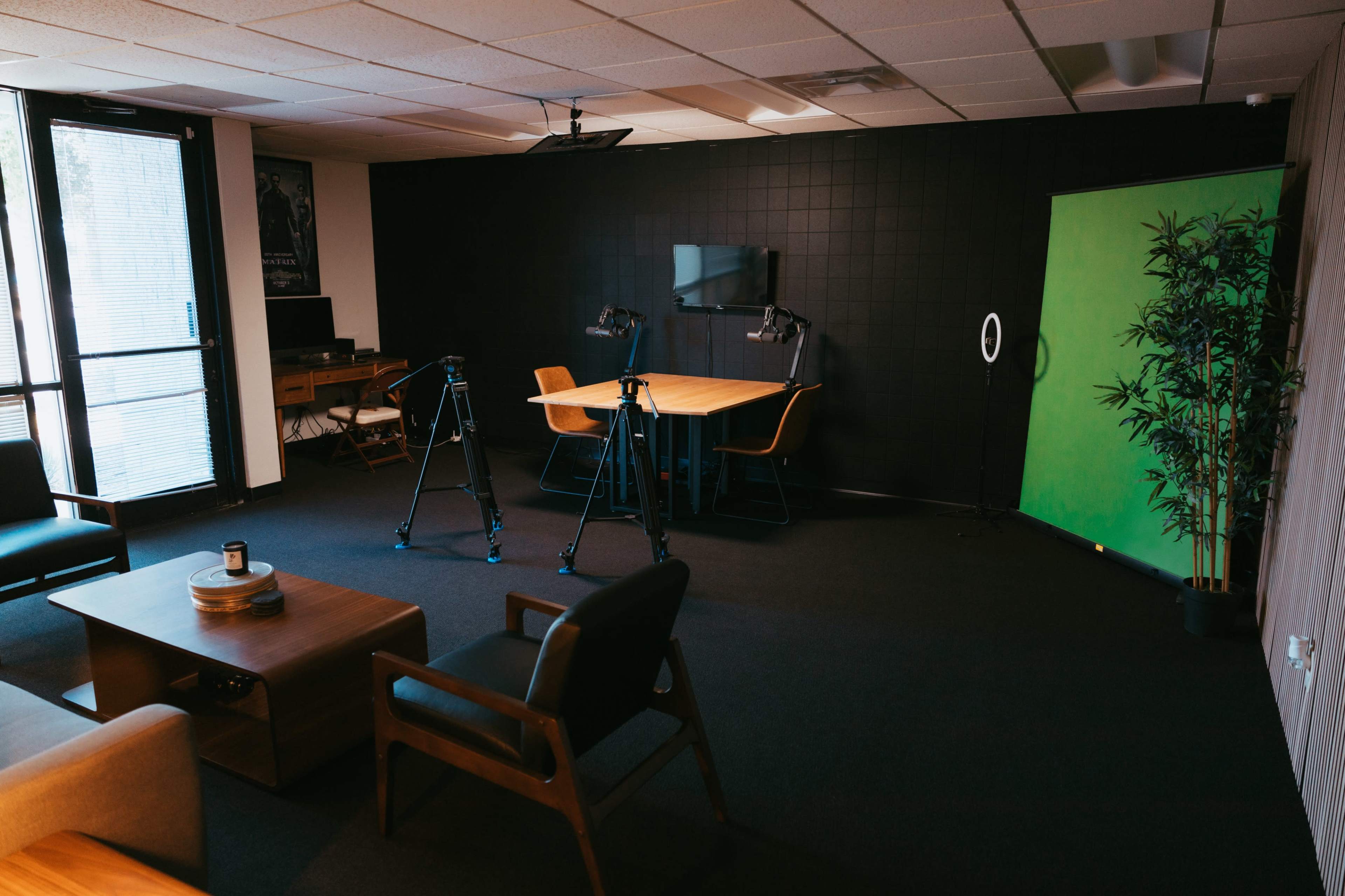 A dimly lit office space features a large wooden table surrounded by chairs, tripods positioned for filming, and a green backdrop.