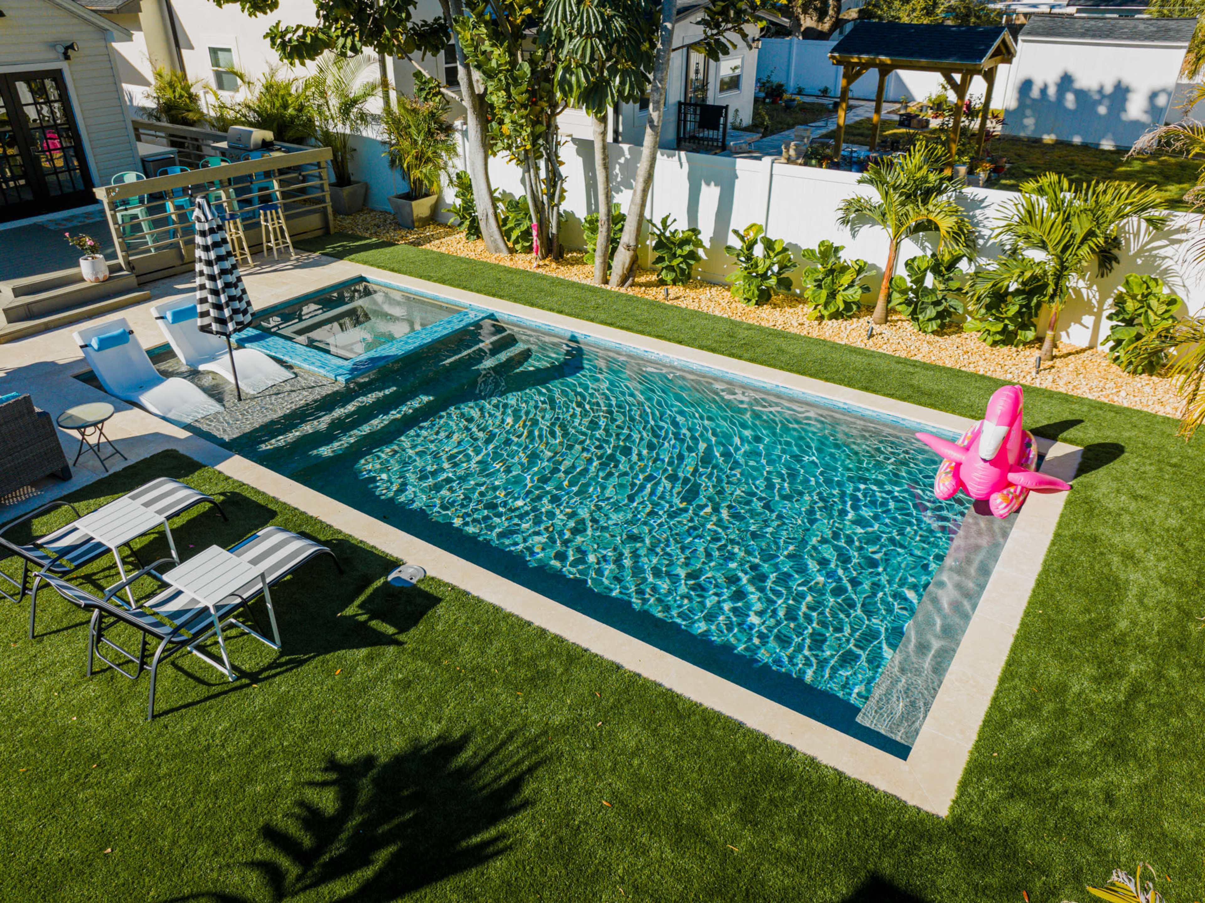 A modern swimming pool surrounded by lounge chairs and a pink inflatable flamingo, with tropical landscaping in the background.