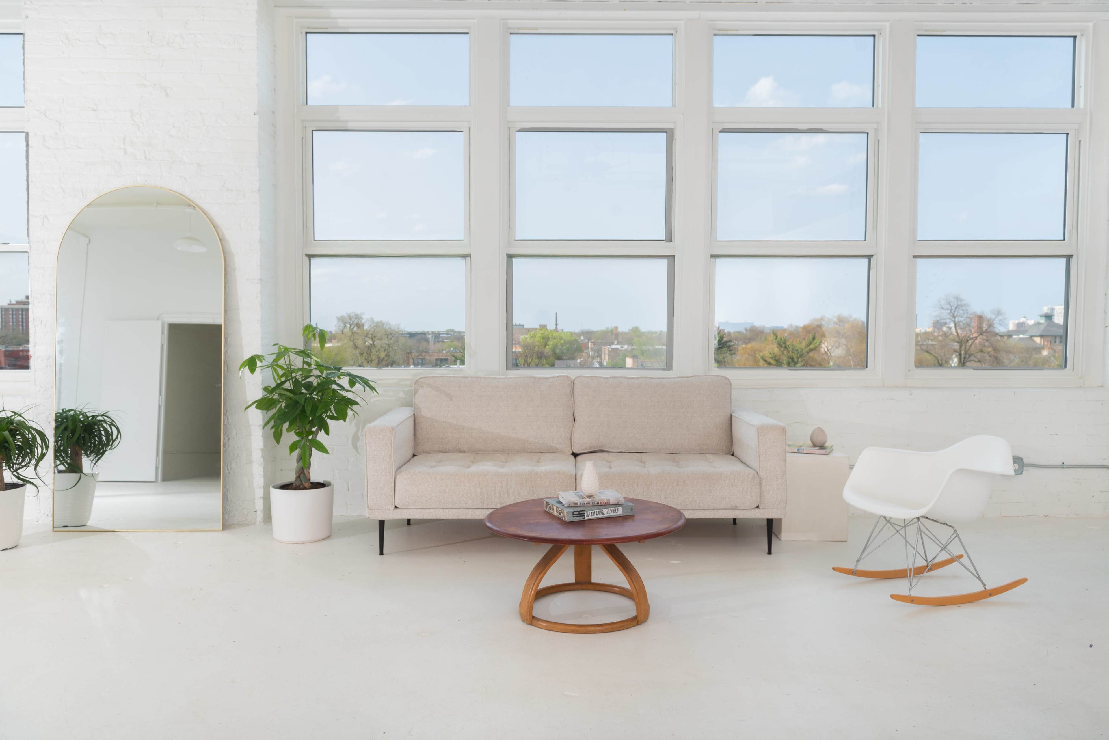 A light-filled room features a beige sofa, a wooden coffee table, a rocking chair, and large windows with a view of the outdoors.