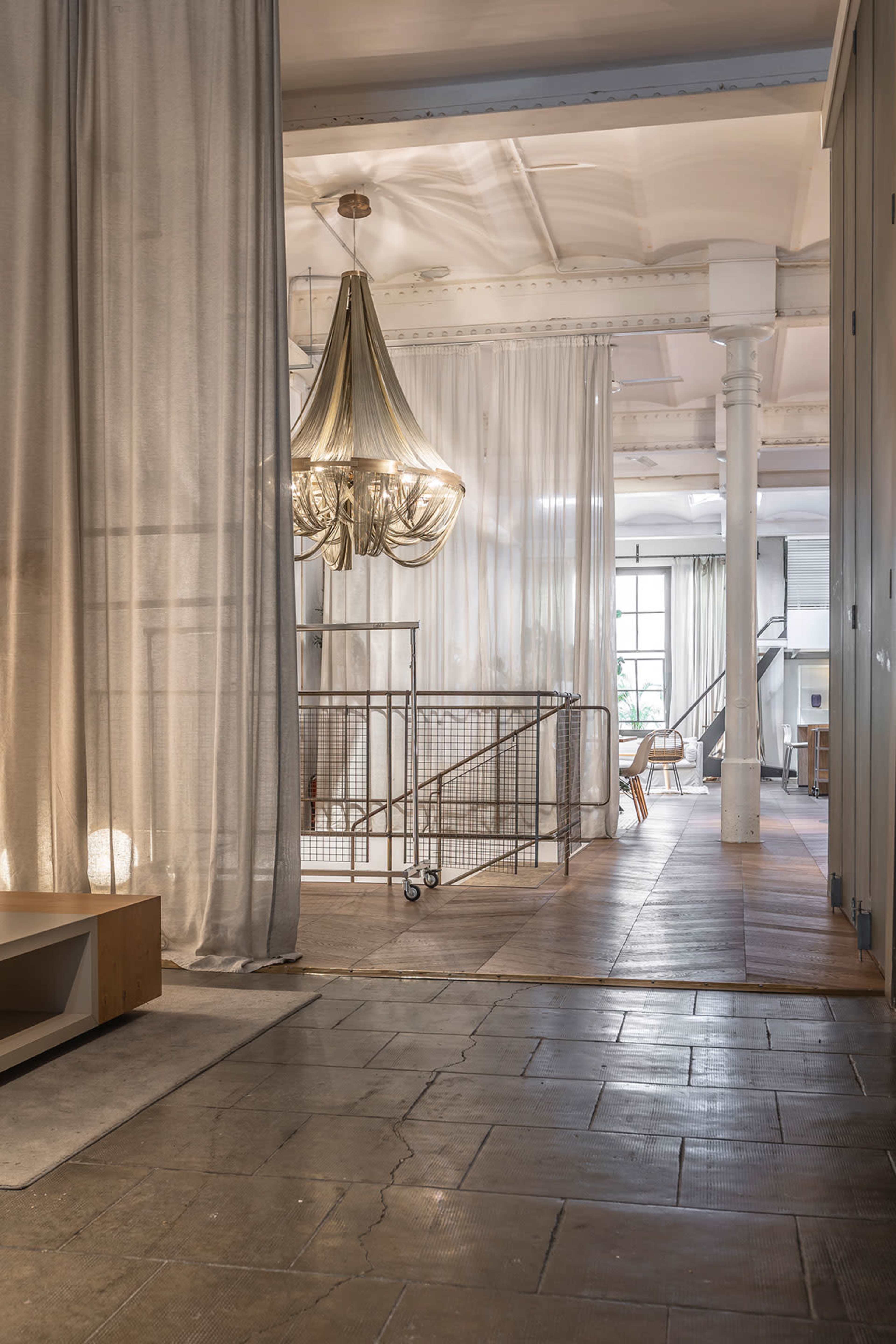 The image shows an interior space featuring a large chandelier, sheer curtains, and a staircase with a metal railing, illuminated by natural light.